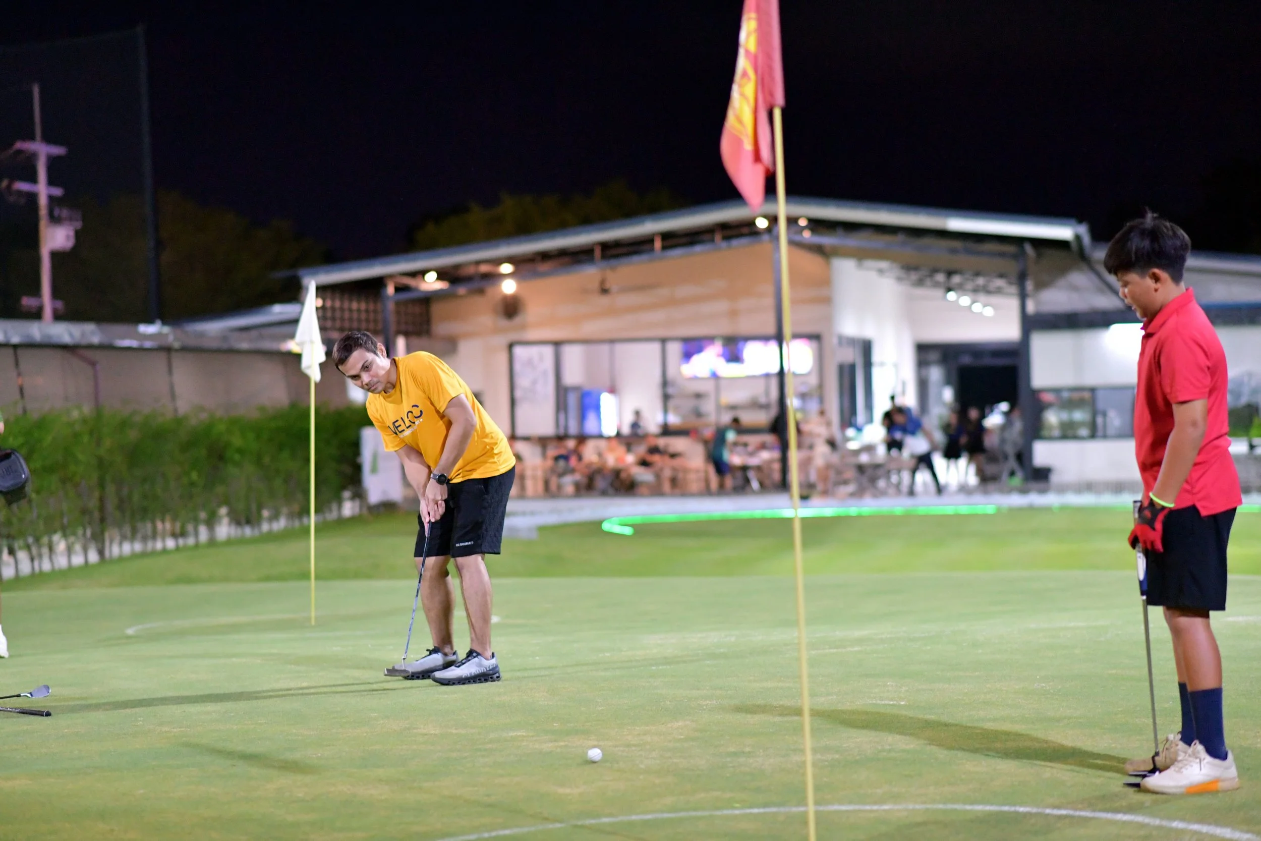 Two young men on a golf green at night, one is putting and the other is holding a golf club, with a clubhouse in the background.