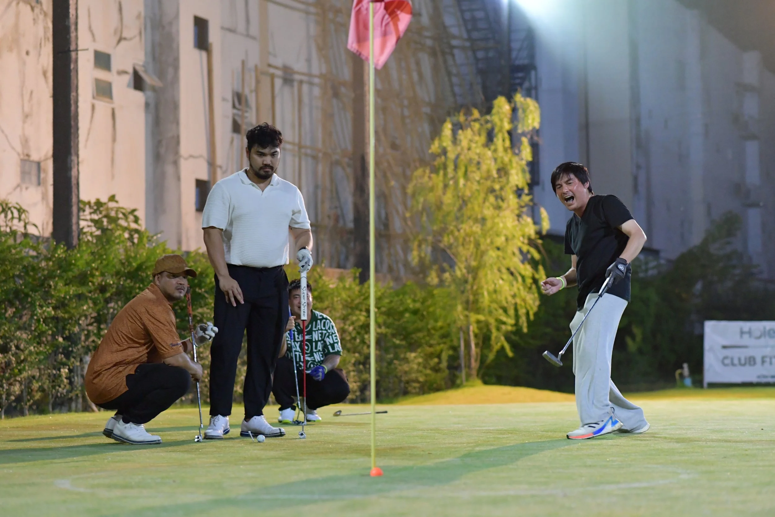 Four men playing golf on a putting green during the evening. One man is putting while the others watch and react, with one squatting and one standing. All are dressed in casual golf attire.