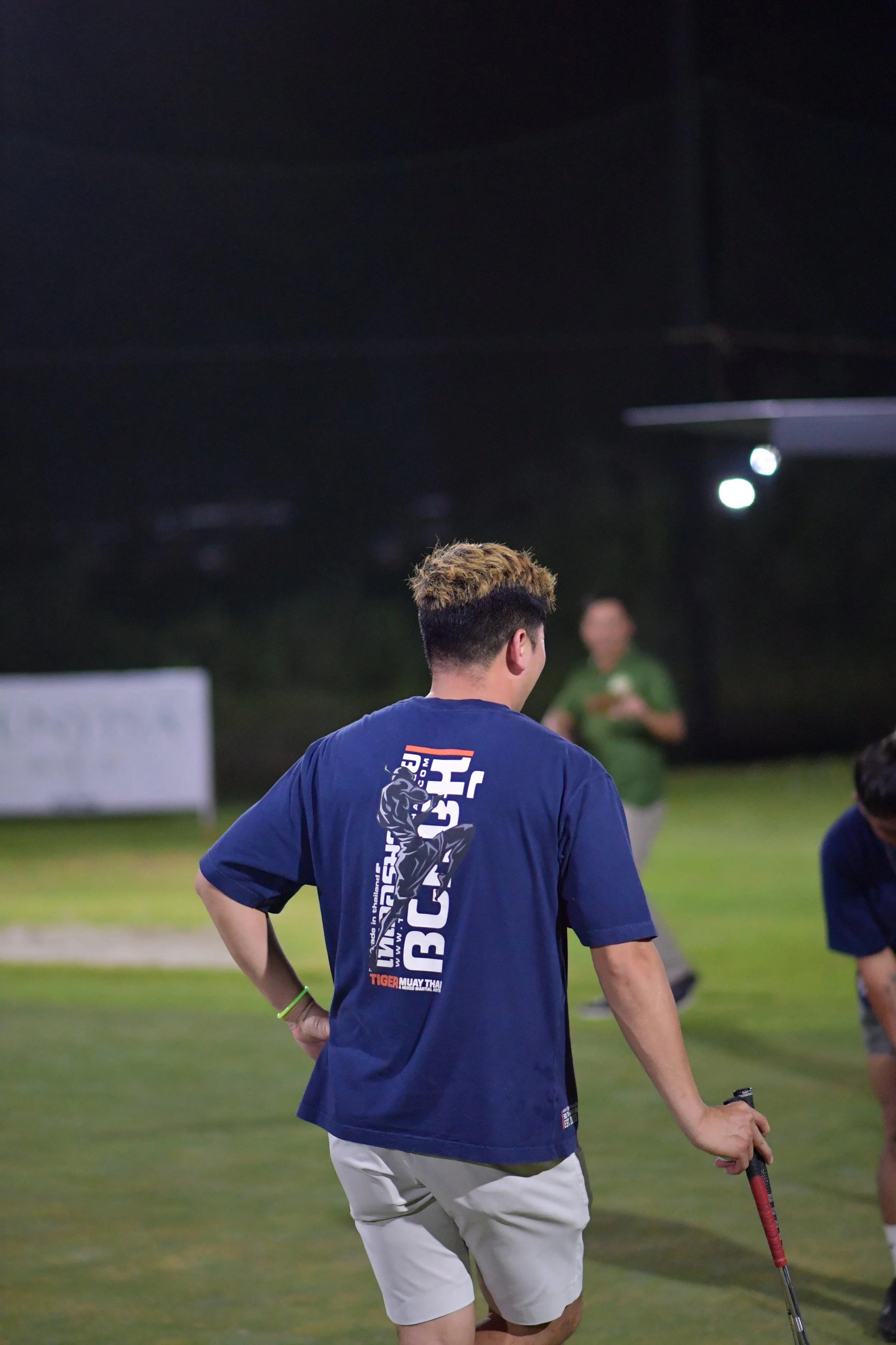 A young man with short hair and light-colored shorts stands on a golf course at night, holding a golf club, with other people in the background.