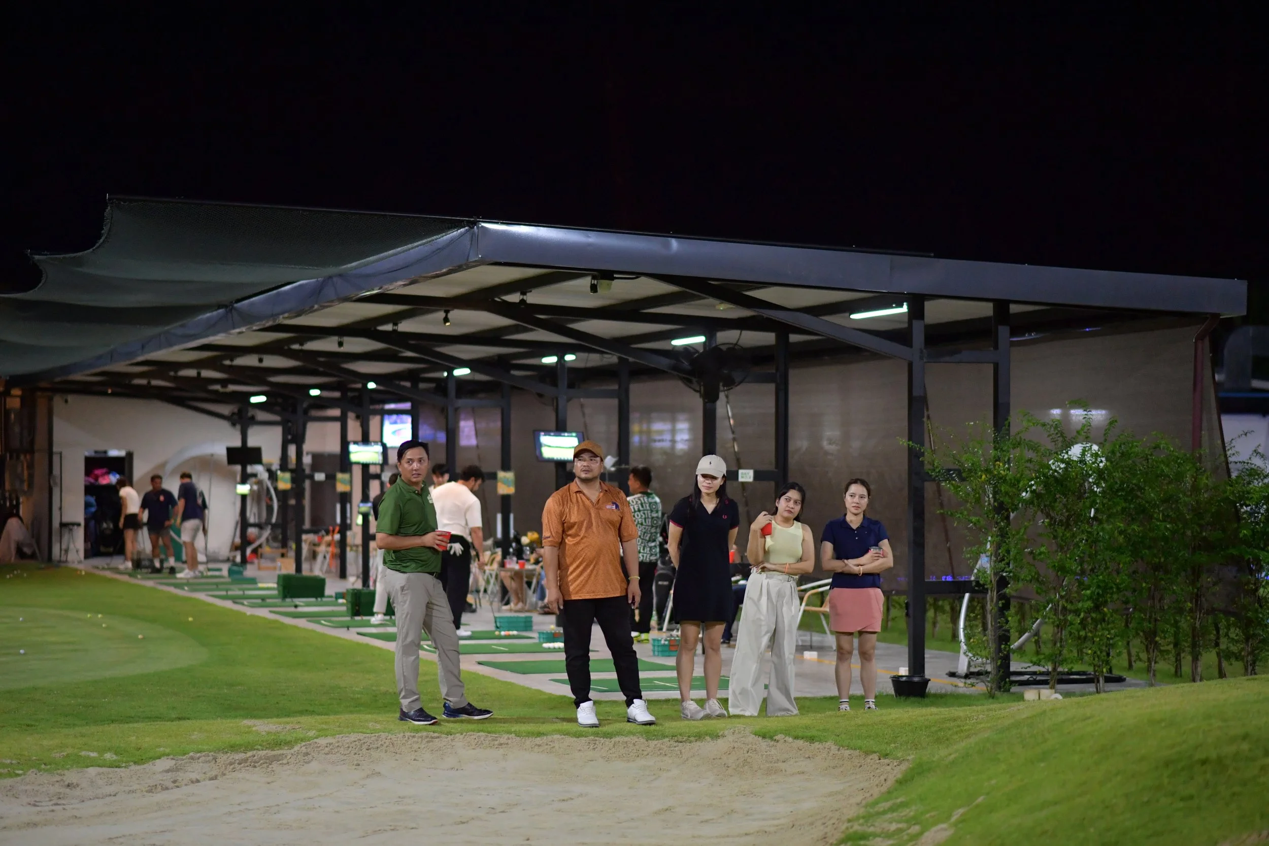 People at a driving range for golf, with some preparing to hit balls and others observing, outside at night under a covered area with digital screens and green grass.