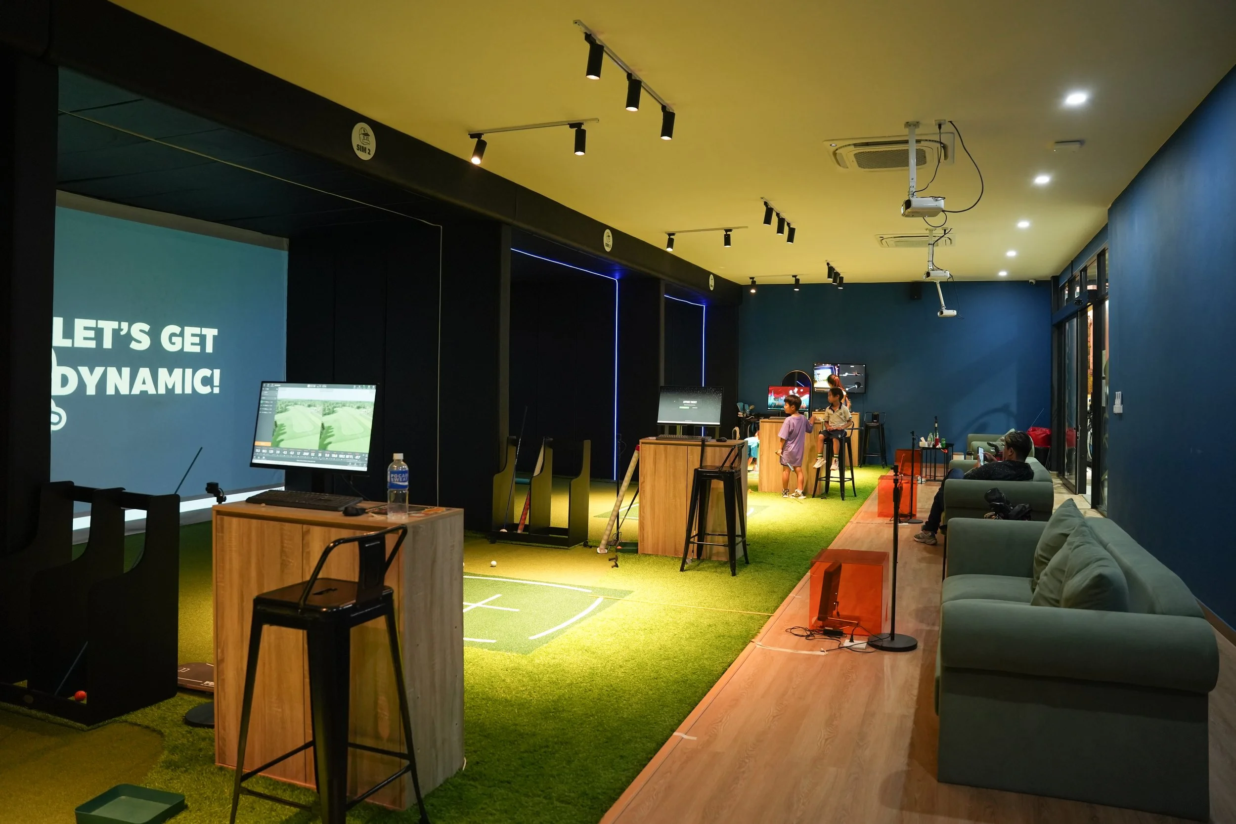 Indoor golf simulation room with a large screen displaying 'LET'S GET DYNAMIC!', several computer monitors, standing and seated people, gaming equipment, and a green turf floor.