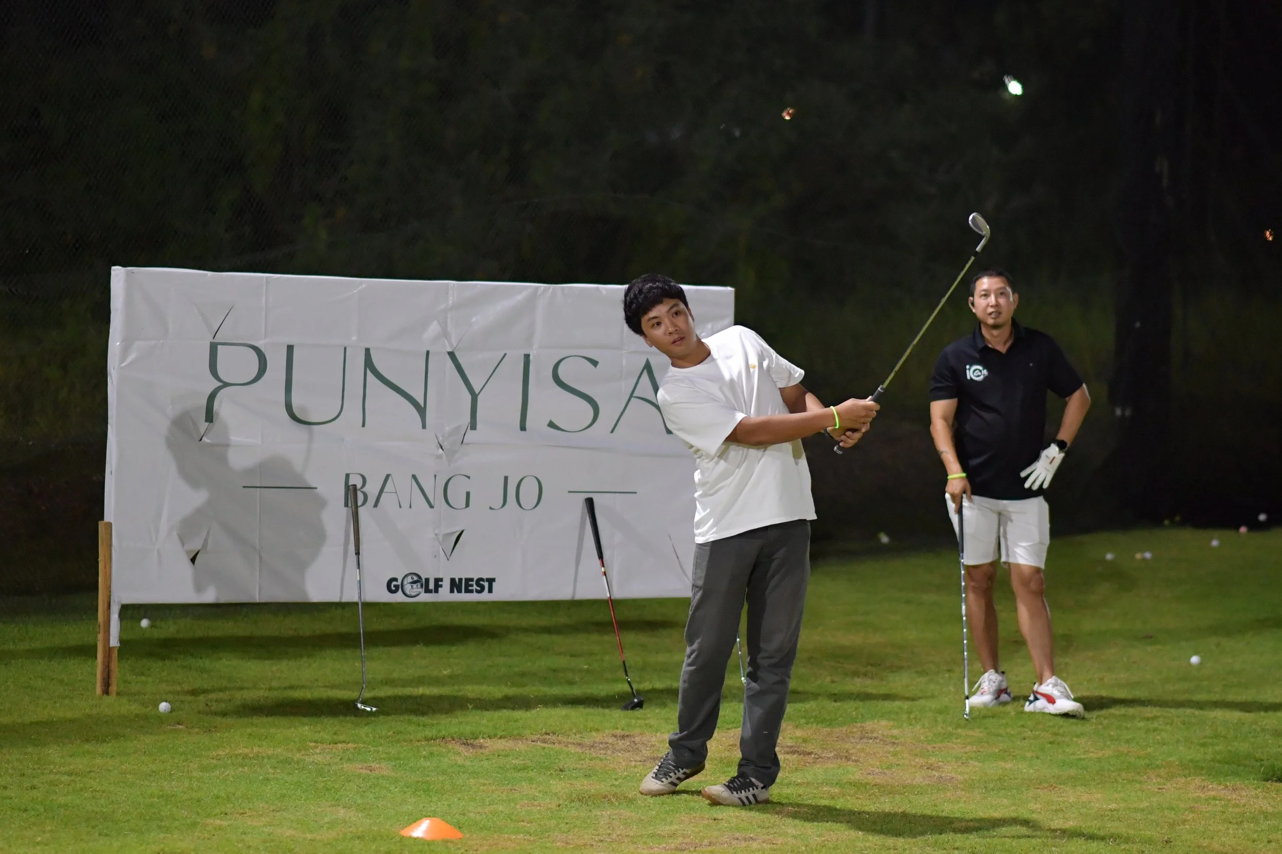 Two men playing golf on a golf course at night; one in white shirt and gray pants, holding a golf club, appears to be taking a shot, the other in black shirt and white shorts, holding a golf club and watching; a sign in the background with the words 
