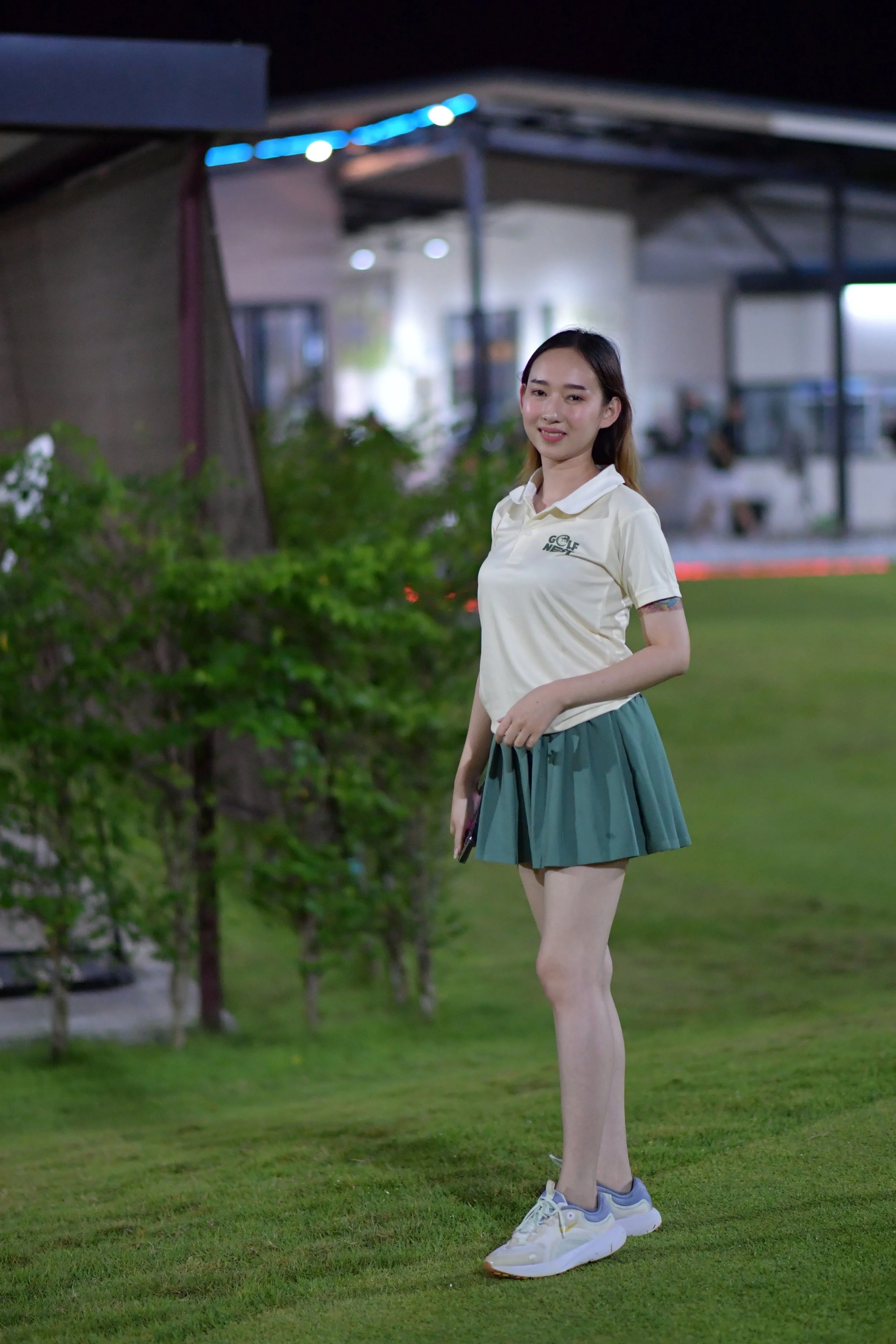 A young woman standing on a lawn at night, wearing a cream polo shirt, teal pleated skirt, and white sneakers, with greenery and a building in the background.