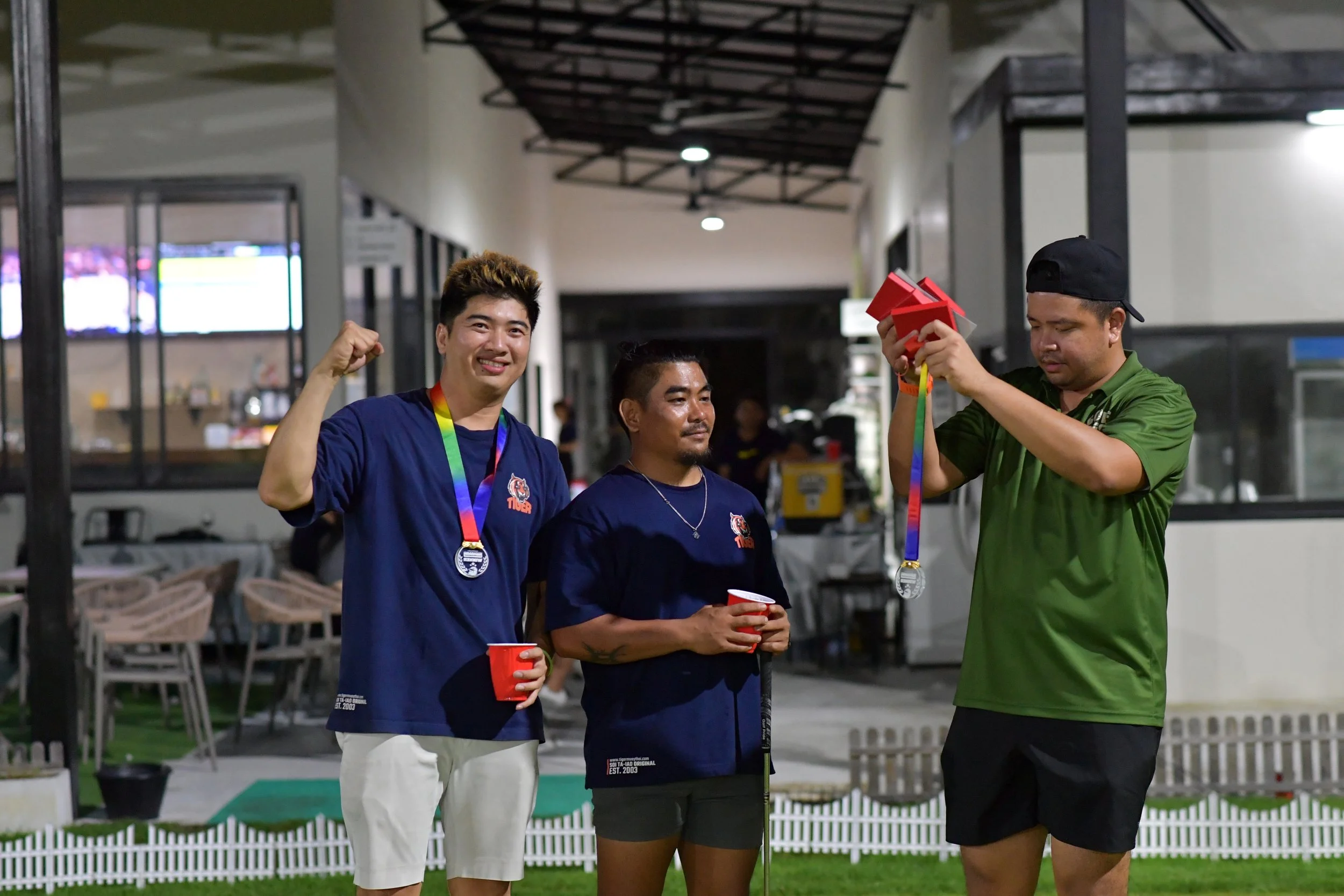 Three people at an awards ceremony outdoors at night. The person on the left and the person in the middle are holding red cups, and the person on the right is putting medals around their necks. The person on the left is smiling and flexing their arm;
