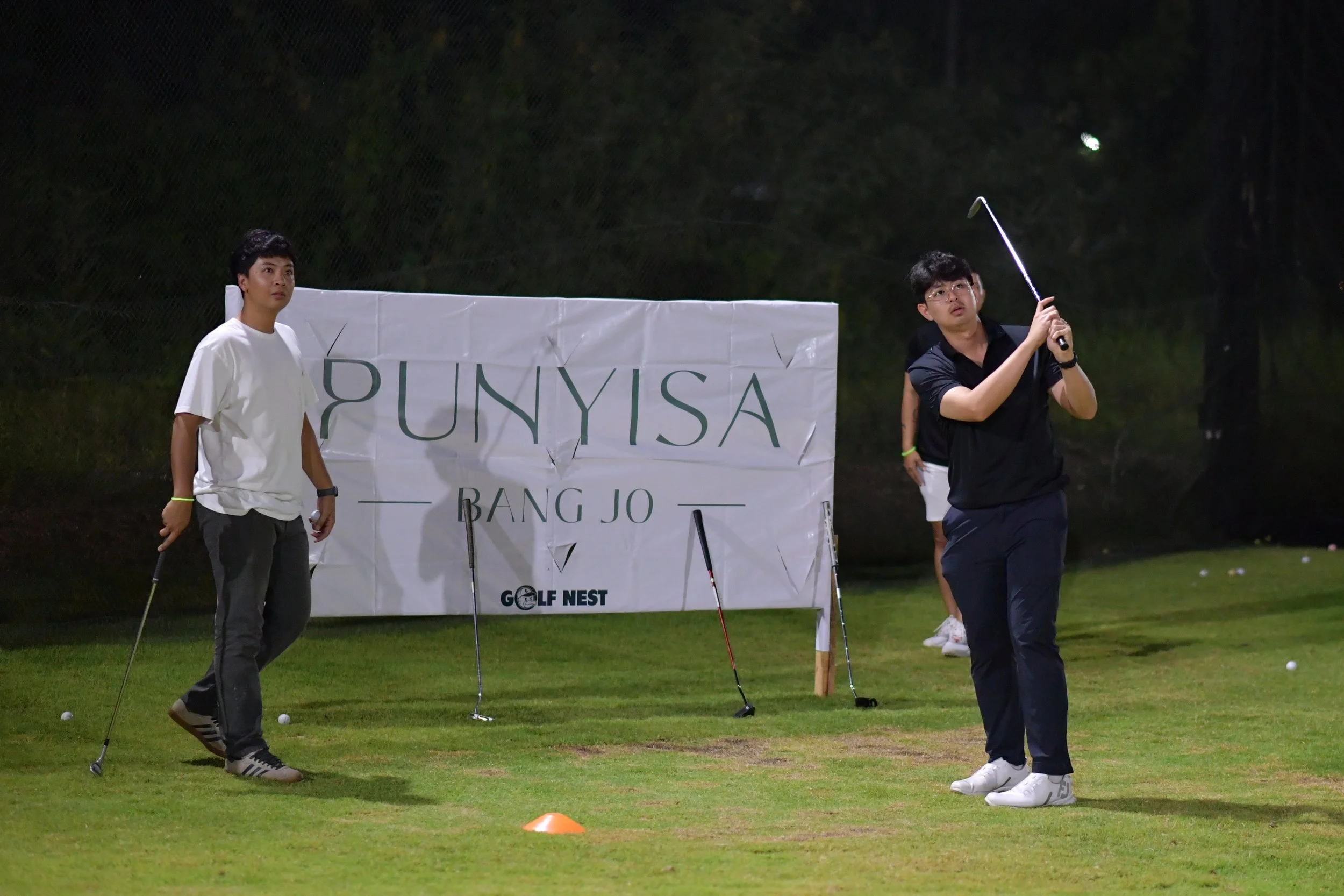 Three young men on a golf course practicing putting, with a sign in the background reading 'PUNYISA BANG JO' and 'GOLF NEST.' The scene appears to be during the evening with artificial lighting.