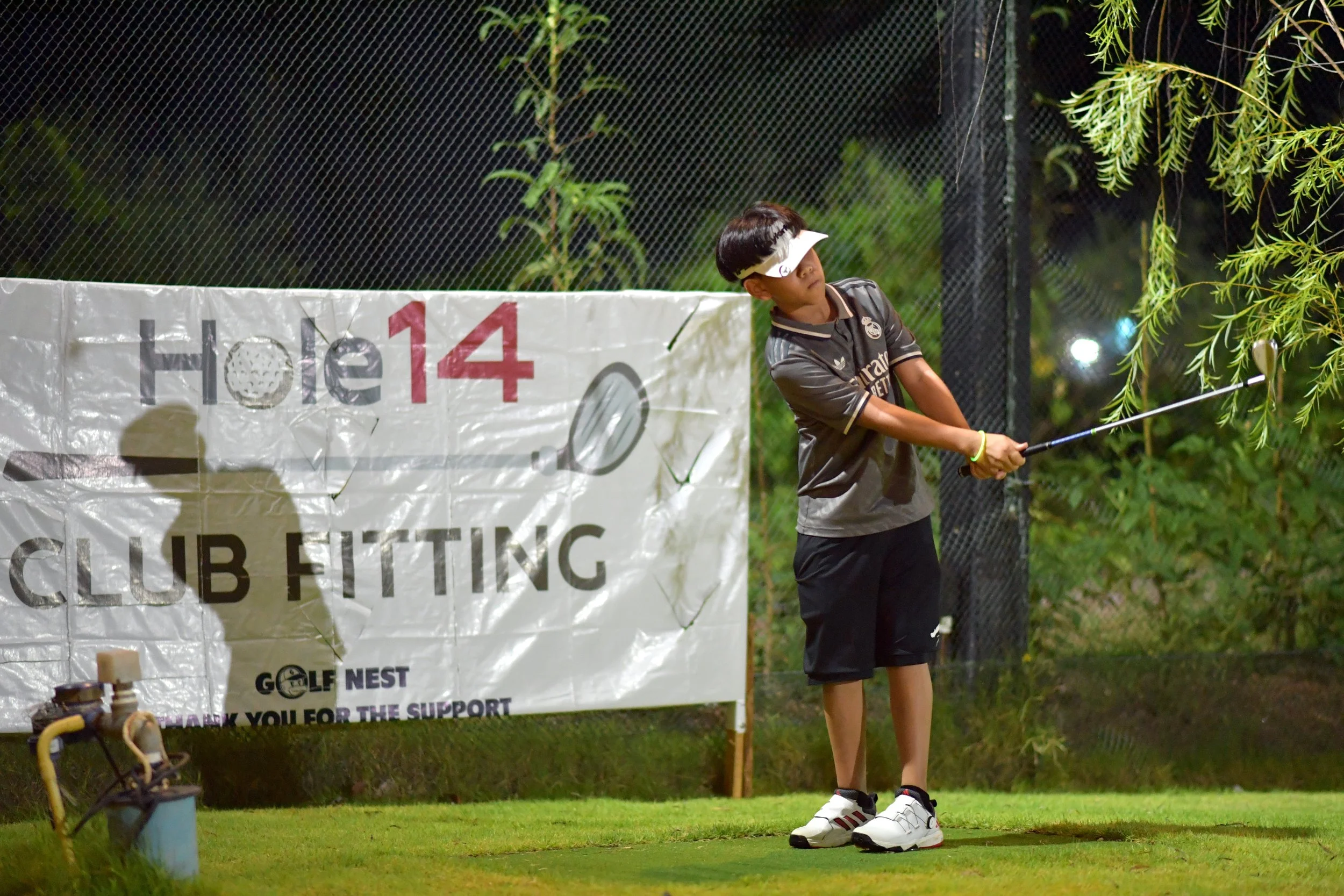 A young boy wearing a black sports uniform and a white visor is swinging a golf club on a golf course at night. There is a large white banner with text and a shadow of the boy cast on it behind him, and greenery surrounds the area.