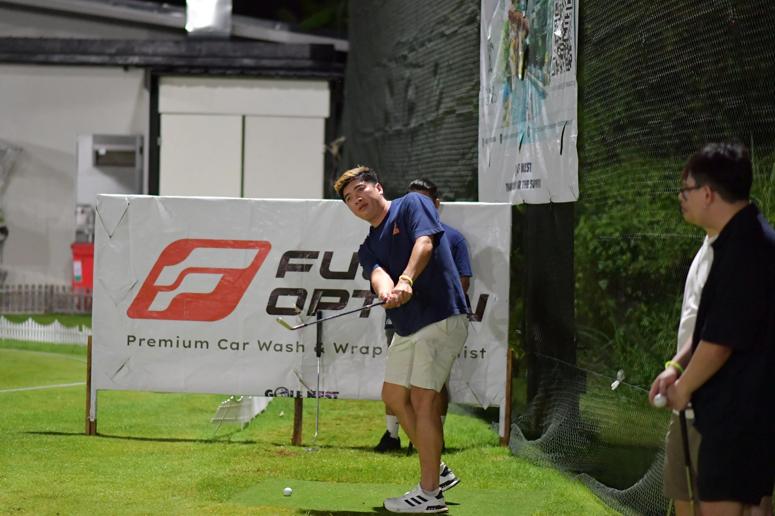 A young man in a navy blue shirt and khaki shorts is preparing to hit a golf ball on a practice mat at a miniature golf course. There is a sign in the background with a logo and text that reads "Fusing Premium Car Wash & Wrap." Two other people are n