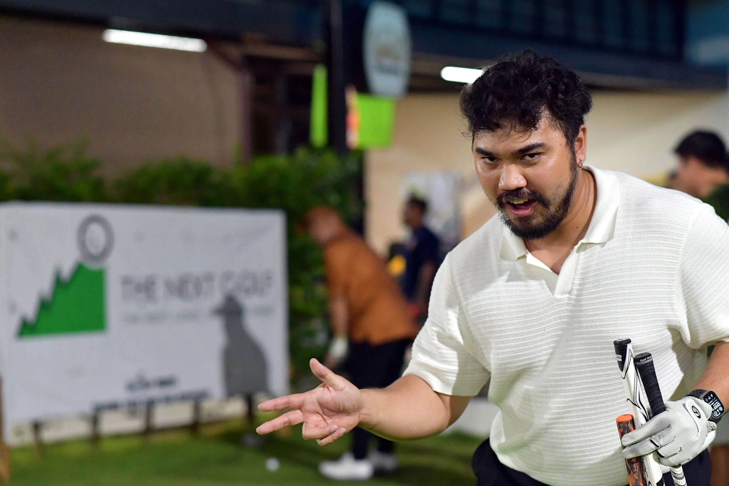 A man with dark curly hair, a beard, and wearing a white polo shirt is holding golf clubs and making a gesture with his hand while standing on a golf course or range. There is a blurred sign and other people in the background.