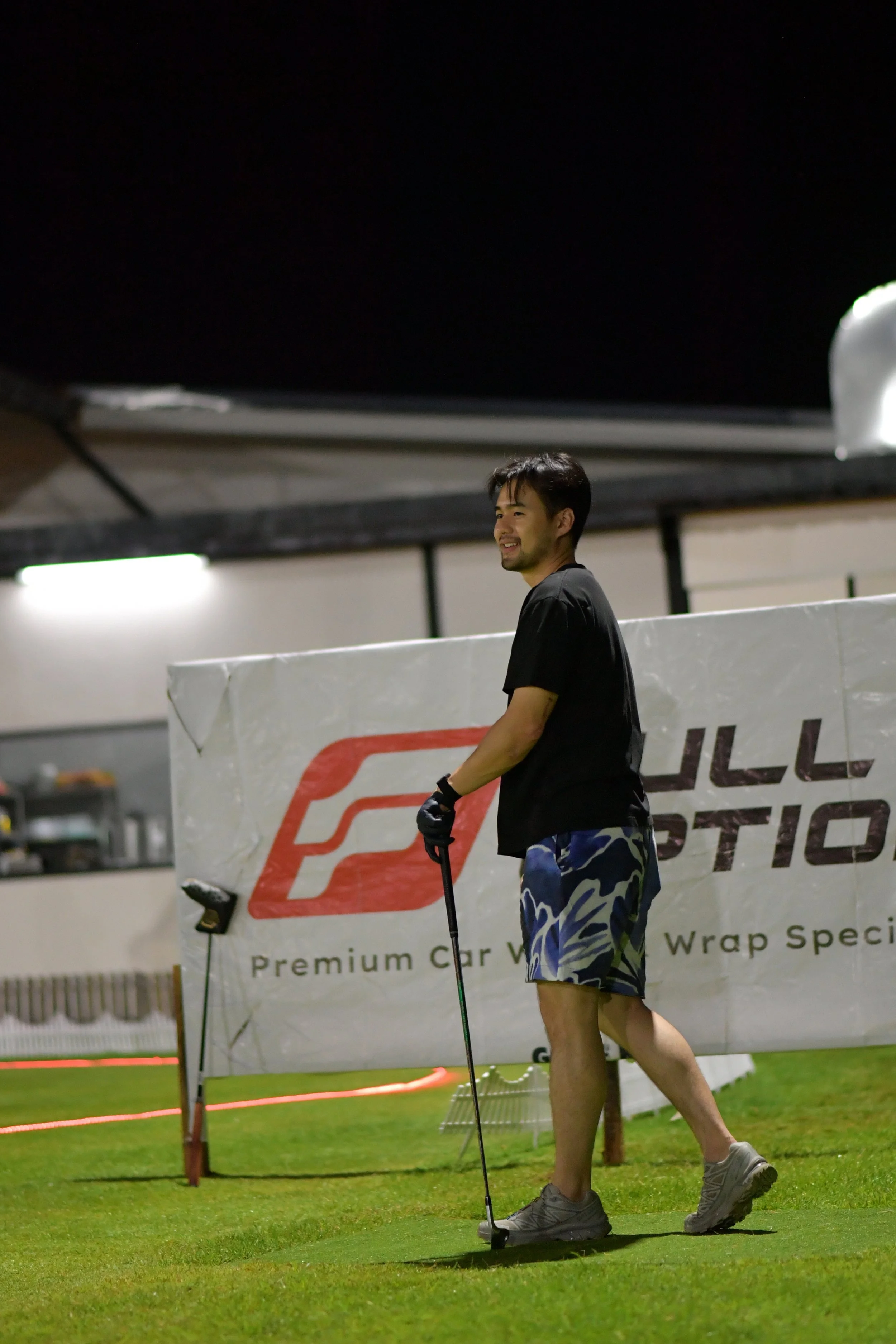 A man smiling on a golf course at night, holding a golf club, with a banner behind him that reads 'Premium Car Wrap Specialist'.