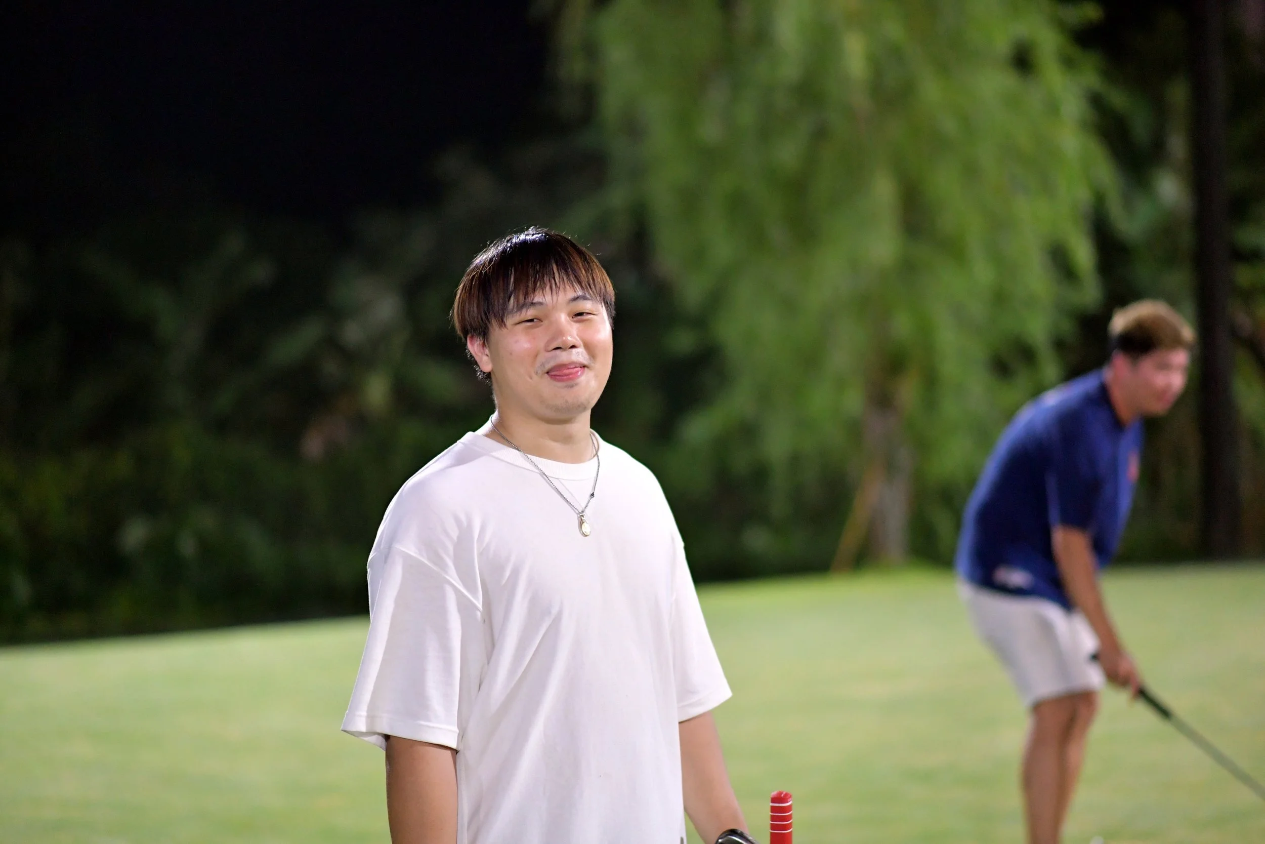 Young man with dark hair, wearing a white t-shirt and a chain necklace, standing on a golf course at night with a smile and tongue sticking out. In the background, another person is preparing to hit a golf ball.
