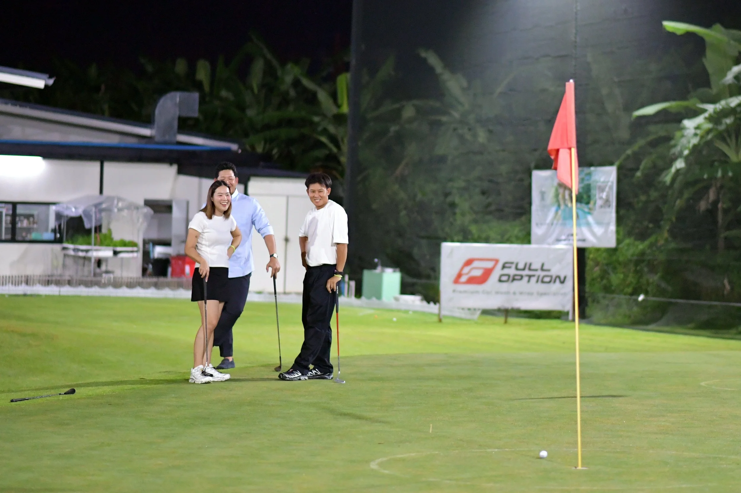 Three people on a golf course at night, standing near a hole with a golf ball nearby. Two men and one woman are smiling and holding golf clubs, with a flagstick and a flag marking the hole.