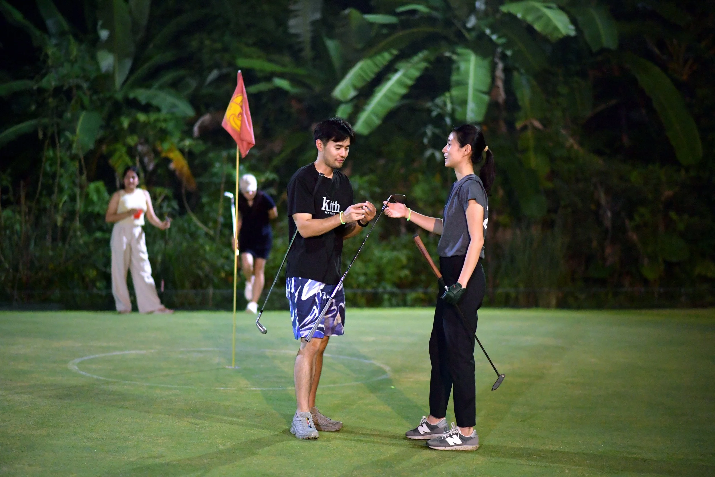 Two people on a golf course talking, holding golf clubs, with a flag and other people in the background.
