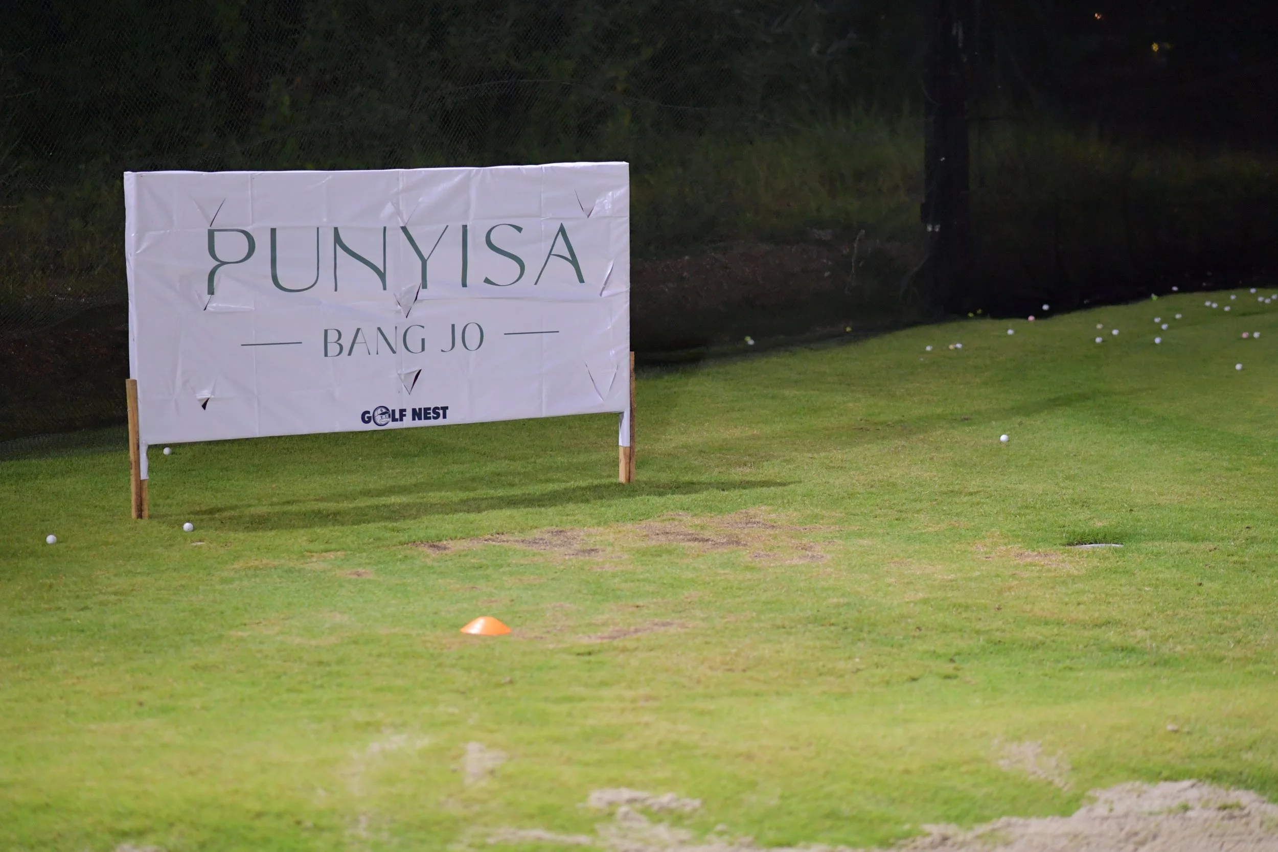 Golf practice area with a large sign reading "PUNYISA BANG JO" and golf balls scattered on the grass, at night.