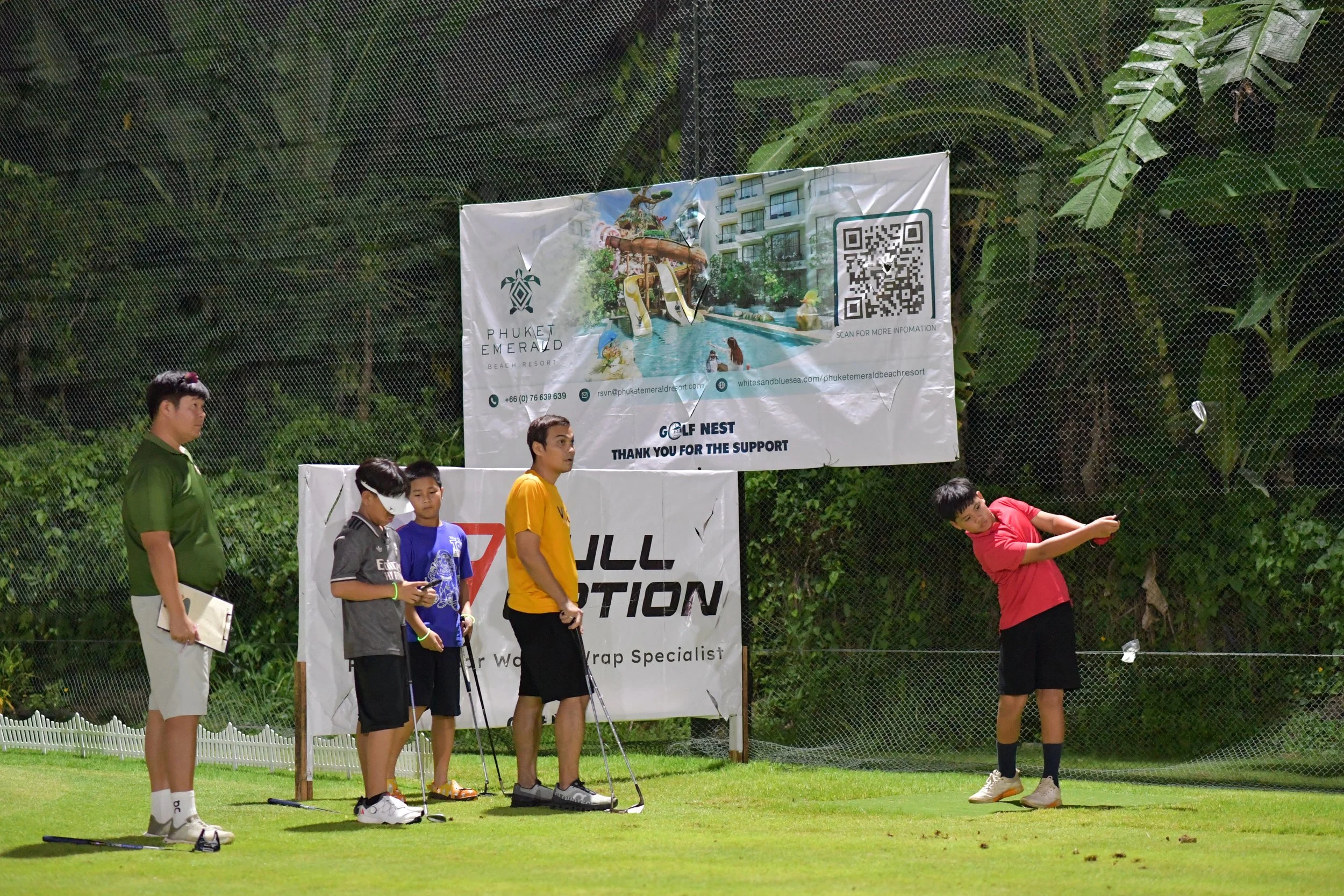A group of people, mostly children, standing on a golf course near a net, with one child swinging a golf club. There are two adults and three children watching, some holding golf clubs. Behind them, there are banners advertising Phuket Emerald Beach 