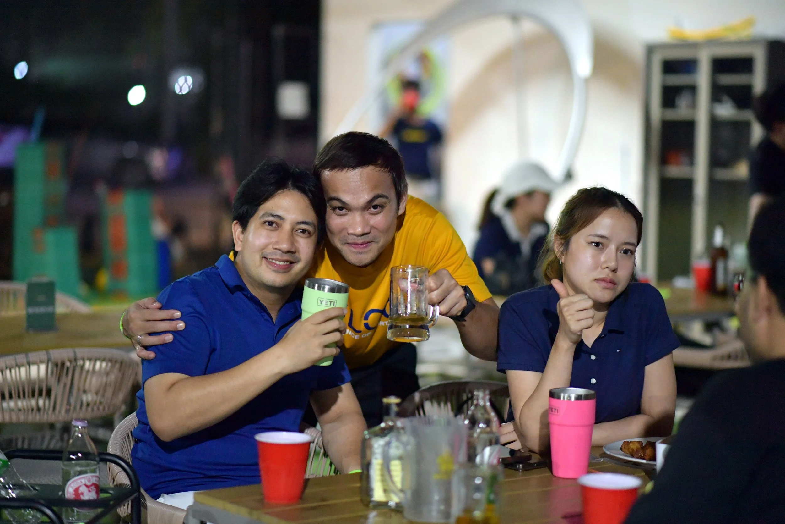 Three friends sitting at a table enjoying drinks and food at a restaurant or bar, with two men holding drinks and a woman with a pink cup, in a casual setting.