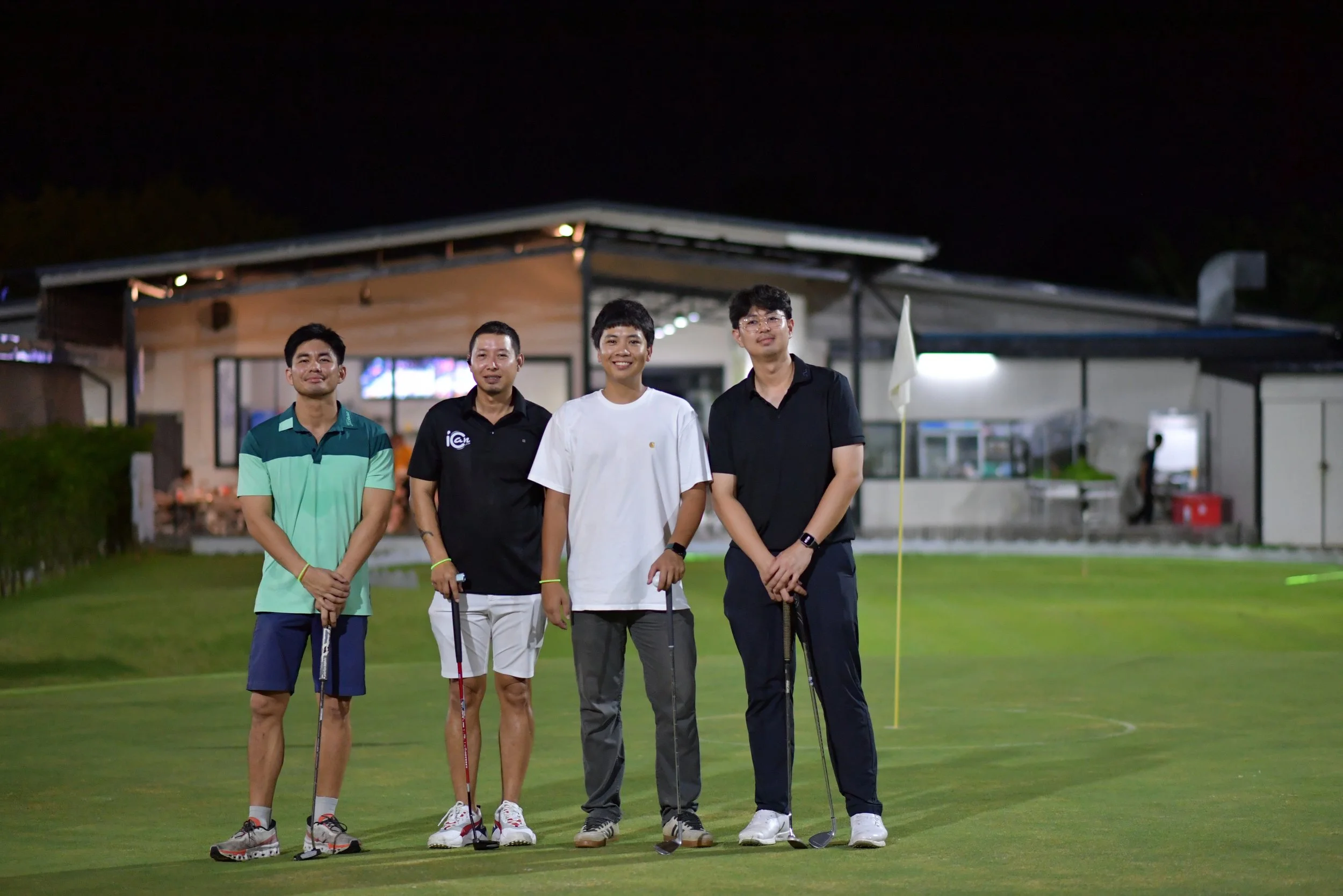 Four young men standing on a golf course at night, holding golf clubs, with a building and a flag in the background.