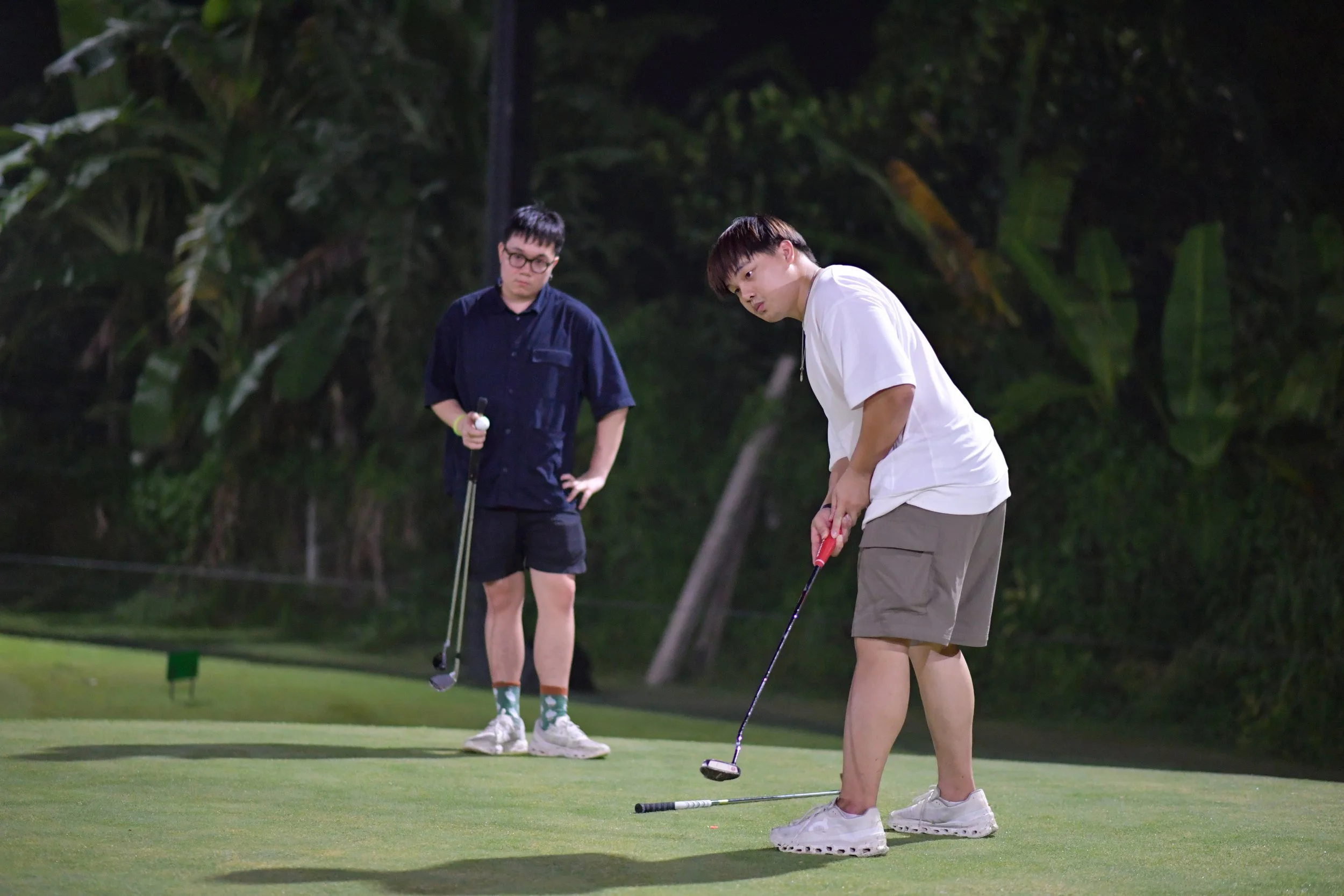 Two young men playing golf on a golf course at night, with one about to putt and the other watching, surrounded by trees.