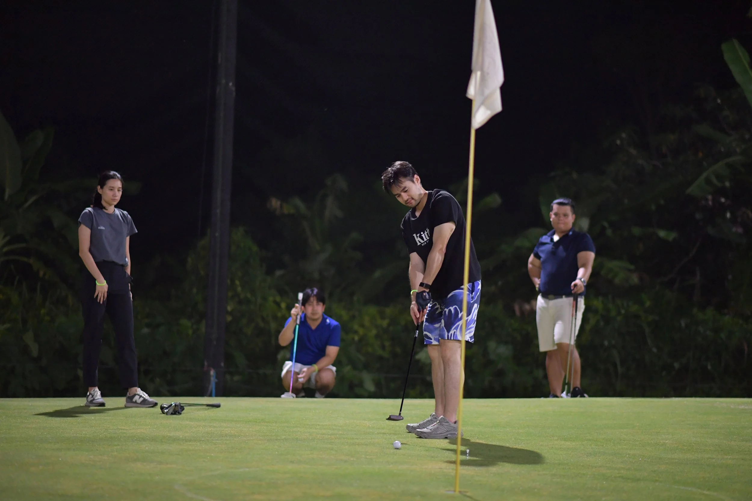 A group of people playing or practicing golf on a putting green at night. One man is preparing to putt, while three others stand or kneel nearby, observing.