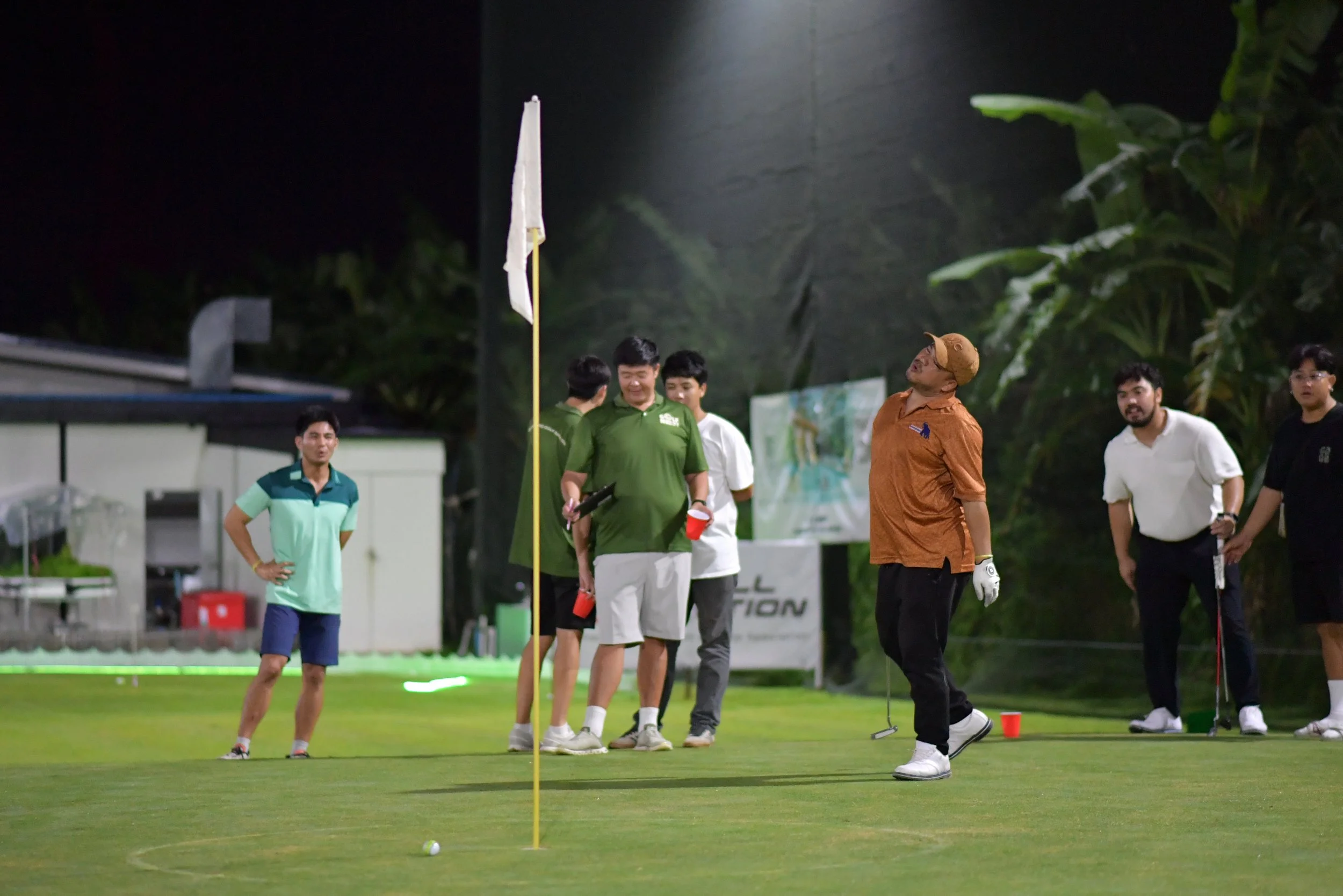 Group of people on a golf course, with a man in an orange shirt and cap taking a shot near the hole, while others watch and chat, some holding golf clubs, during what appears to be a casual golf game at night.