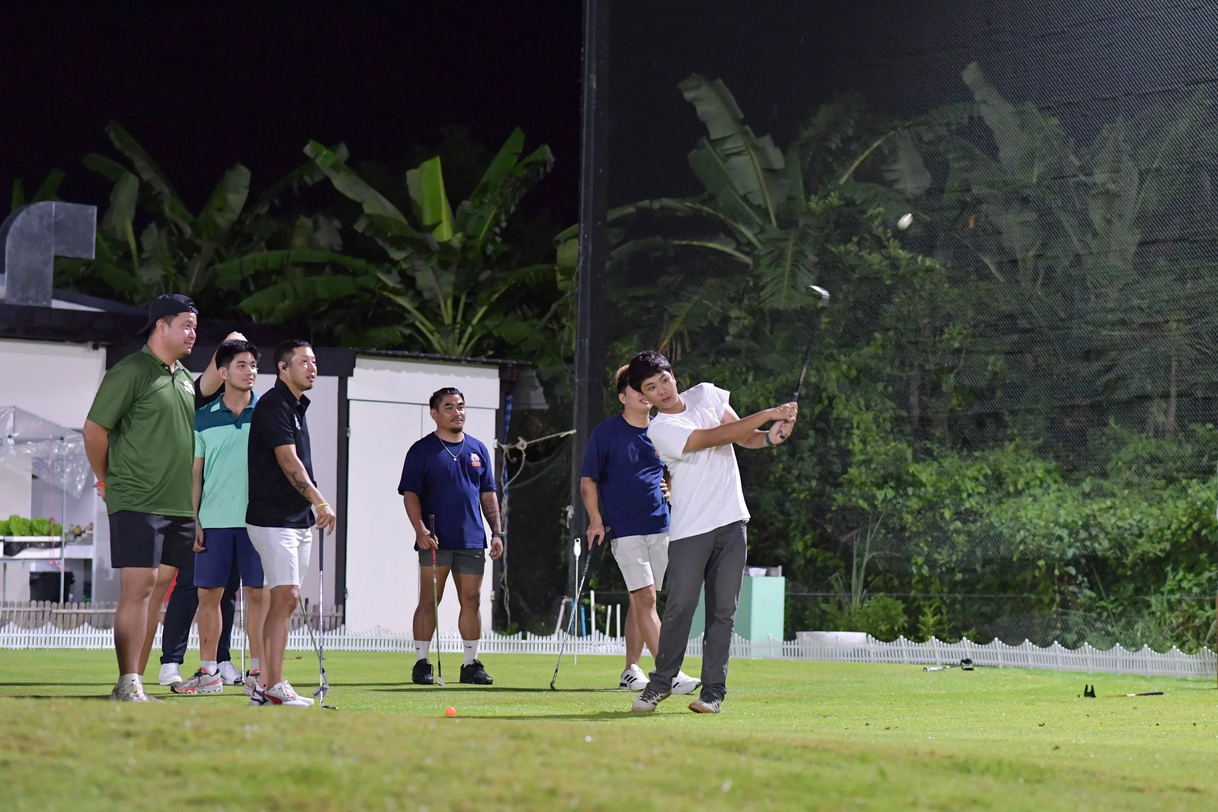 Group of men playing night golf on a course, with one man swinging a golf club while others watch, surrounded by greenery and a netted background.