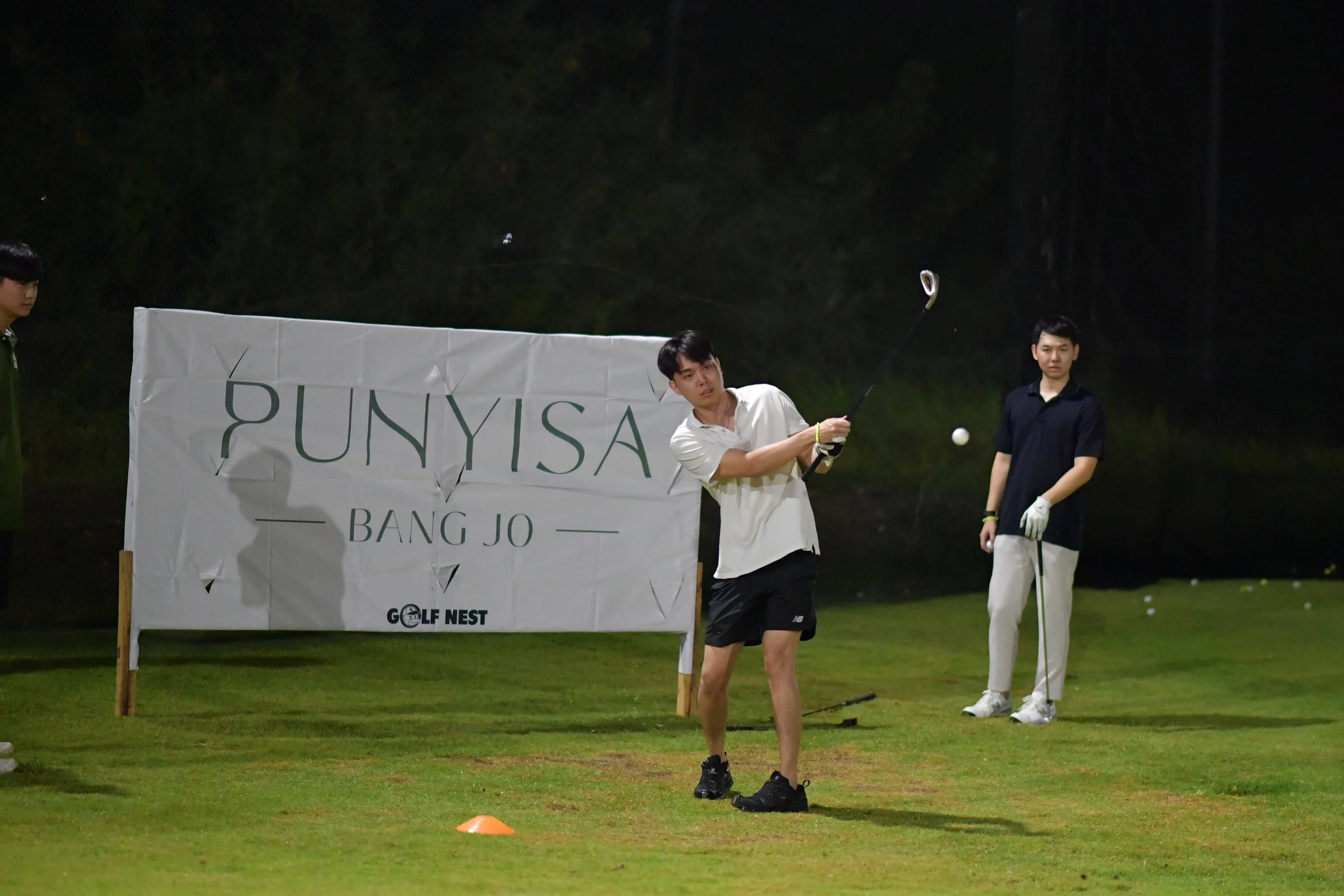 A young man playing golf at night on a well-lit golf course, with a large white sign that reads 'PUNYISA BANG JO GOLF NEST' in the background. Two other young men stand nearby, one watching and the other holding a golf club.