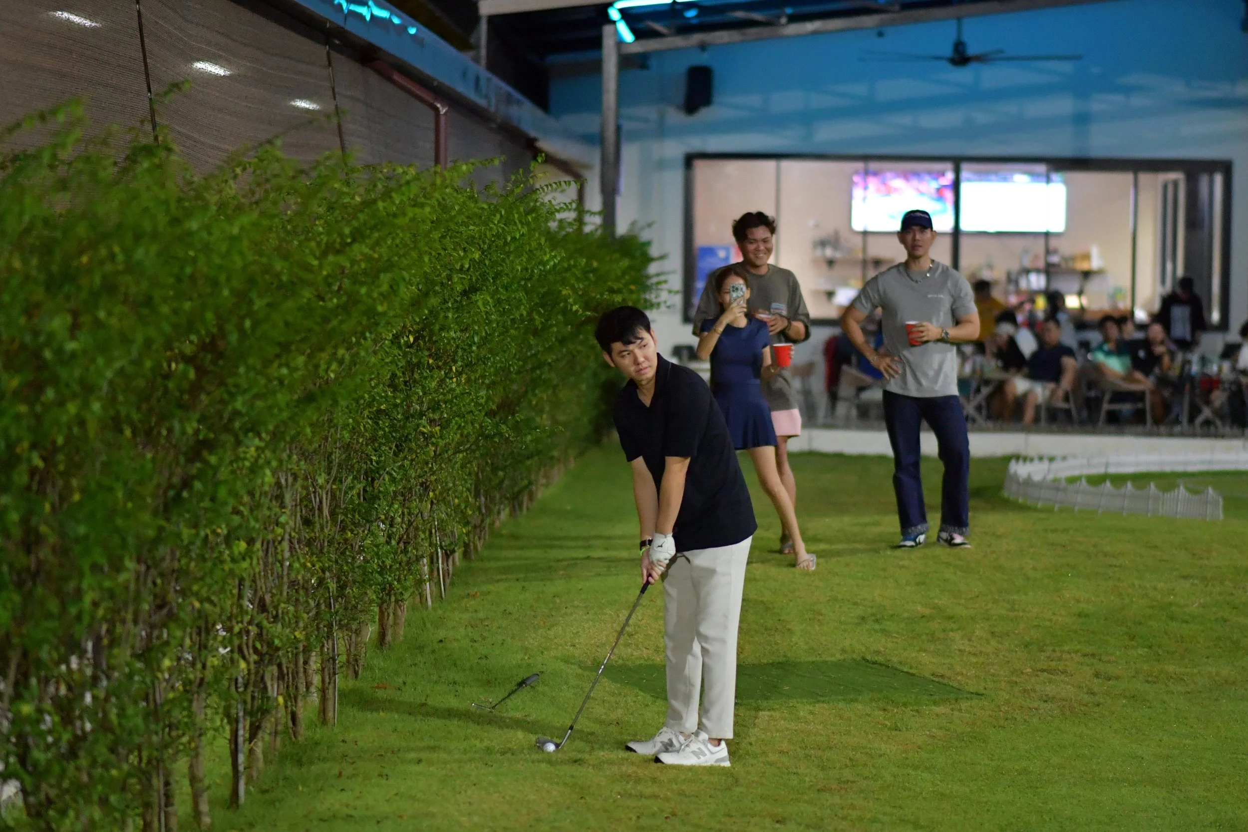 A woman is playing golf, preparing to hit a ball on a grassy patch. Three people are watching her in the background, two holding drinks and one taking a photo, at a golf course or club during the evening.