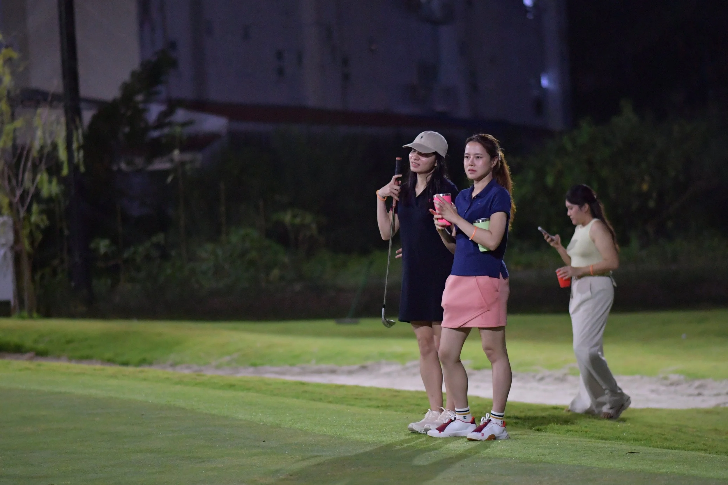 Three women on a golf course at night, two of whom are standing close together looking at their phones, and a third woman in the background also using her phone, with lush trees and a building in the background.