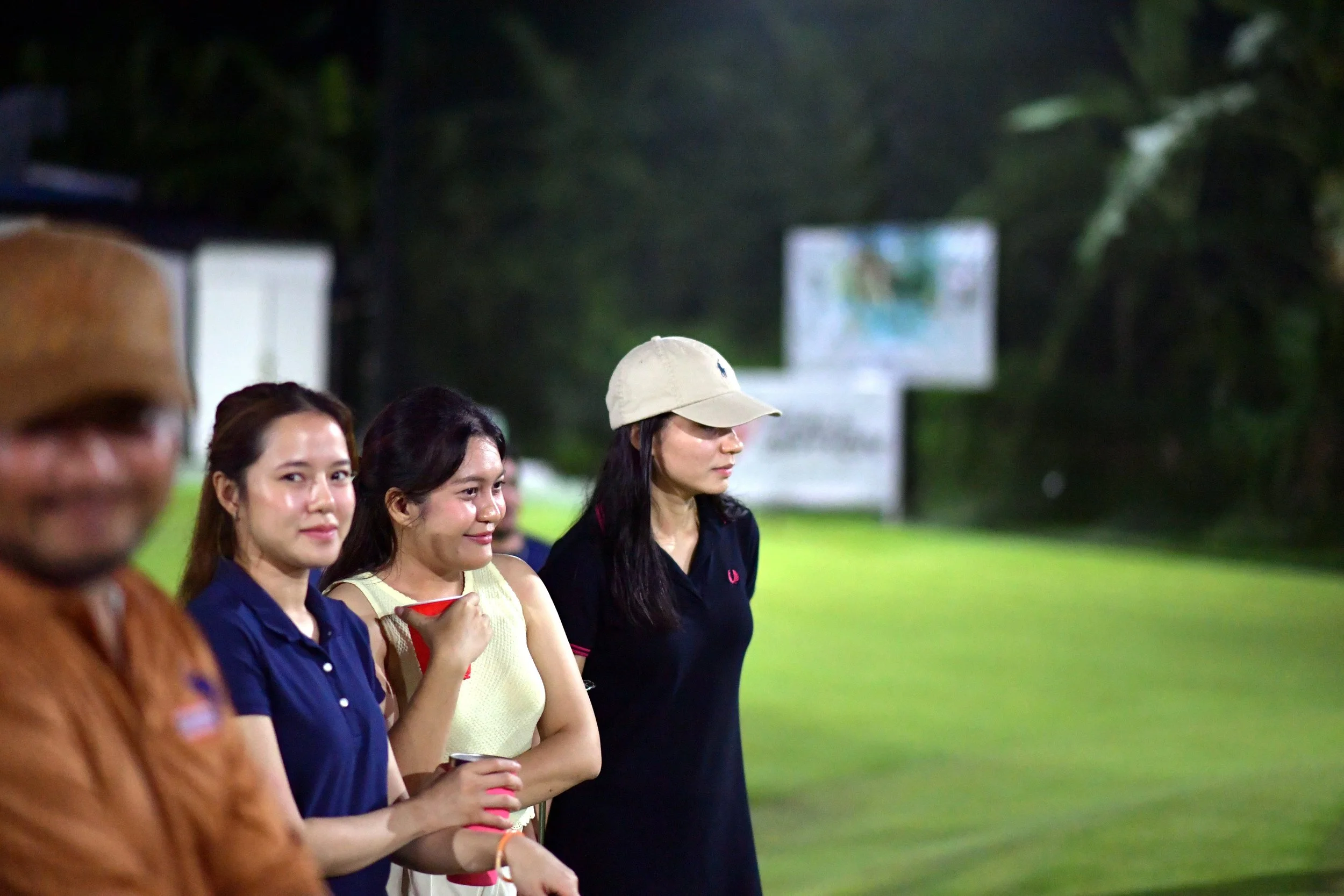 A group of mainly women standing outdoors at night, dressed casually, with a grassy background and blurry signs or billboards in the distance.