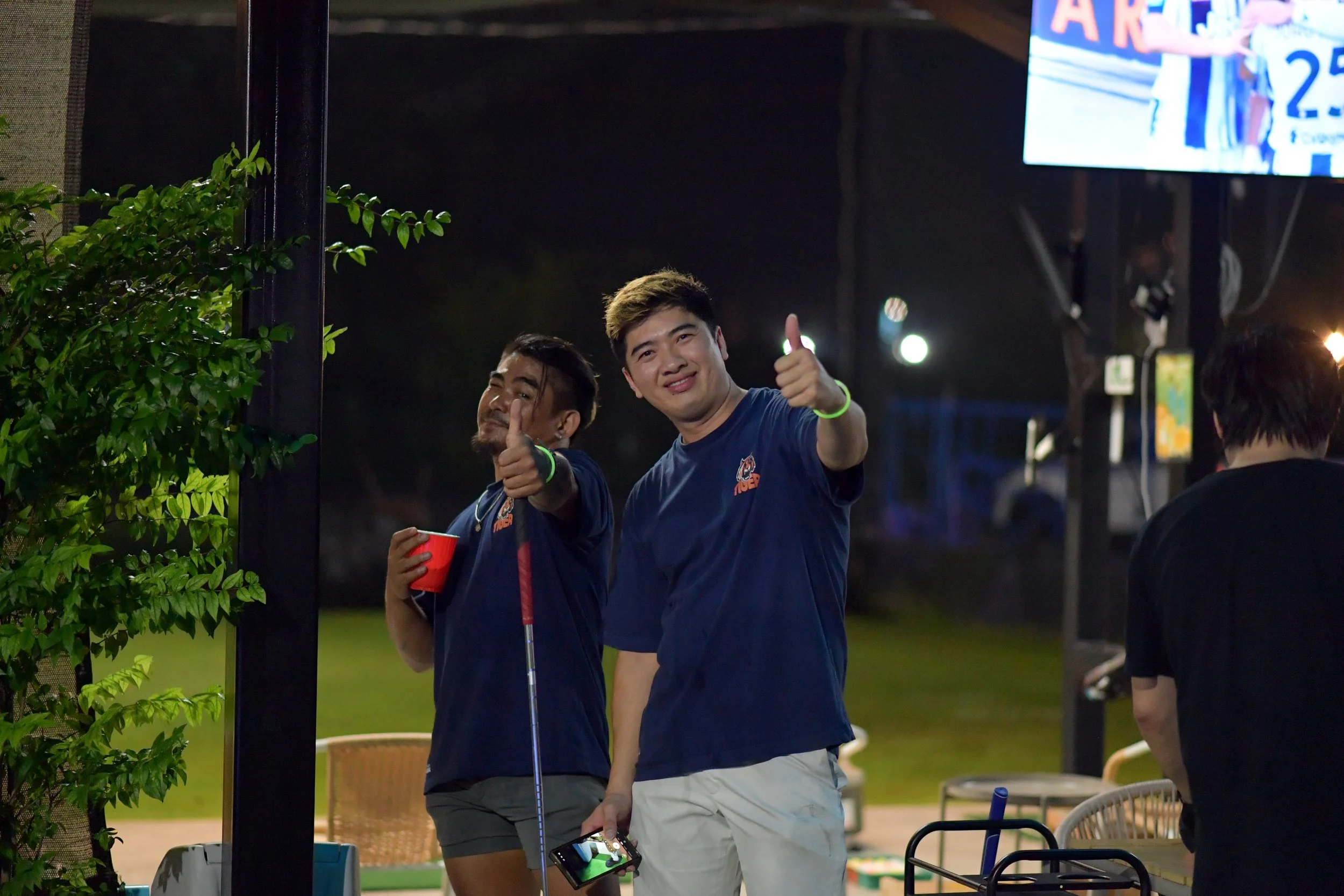 Two men in blue shirts giving thumbs up at an outdoor event at night. One is holding a red cup and a golf club, smiling at the camera. The other is smiling and holding a phone. There is a large screen and some chairs in the background.