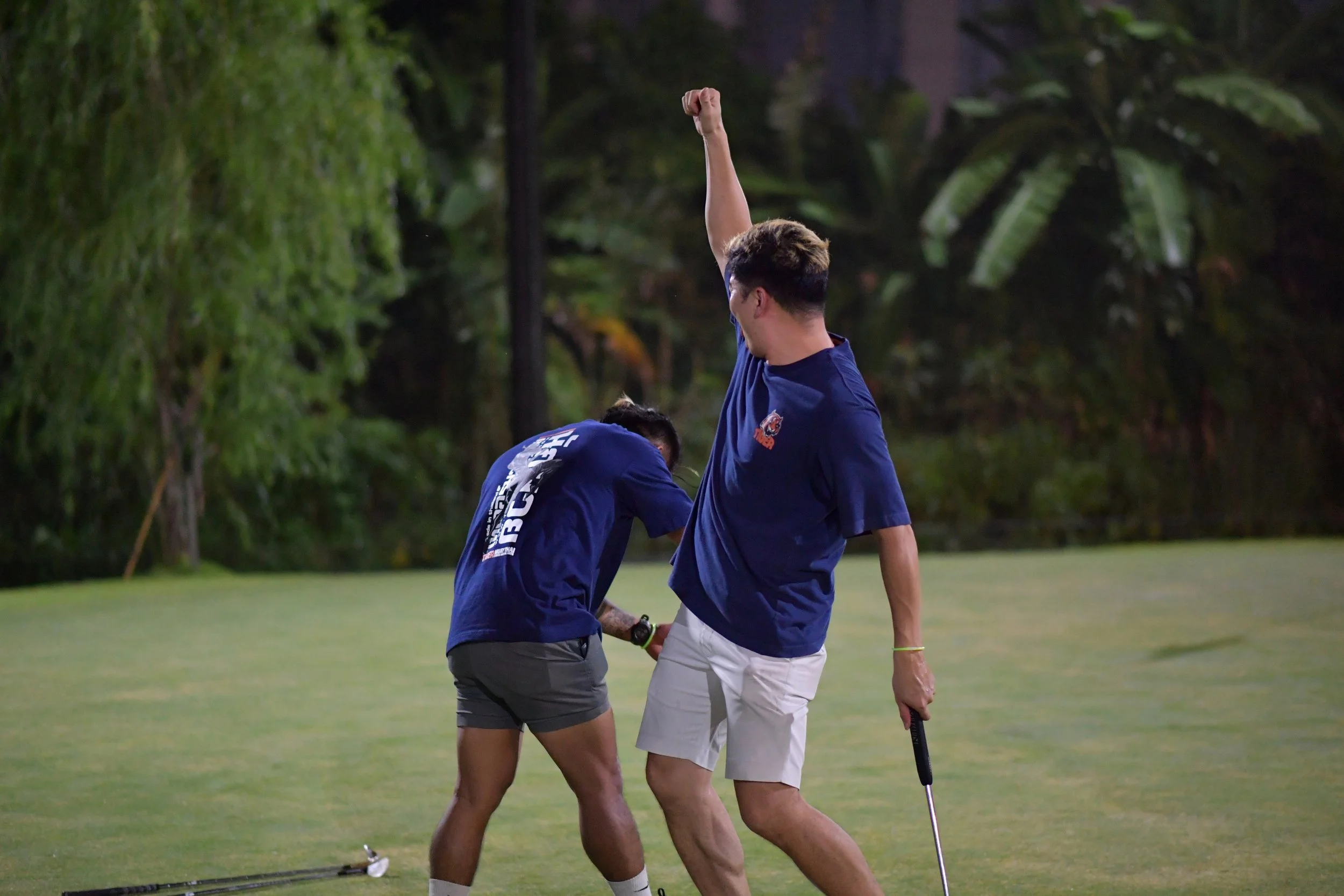 Two men on a golf course celebrating a successful shot or hole. One man raises his fist, while the other bends down.