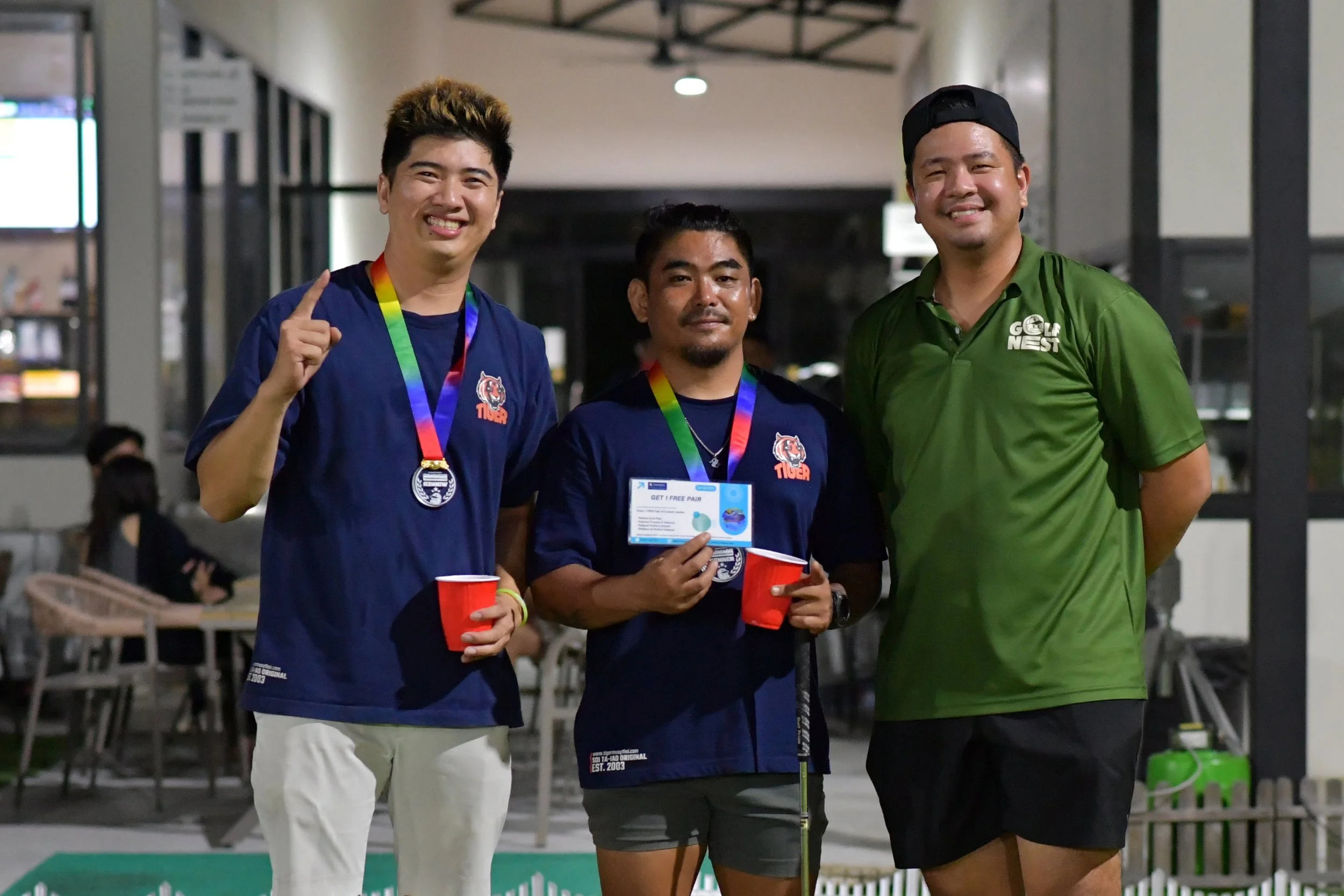Three young men standing together at an indoor event, smiling and posing for the photo. Two are wearing navy blue shirts with a tiger logo, and one is wearing a green shirt with white text. They have medals around their necks and are holding red cups