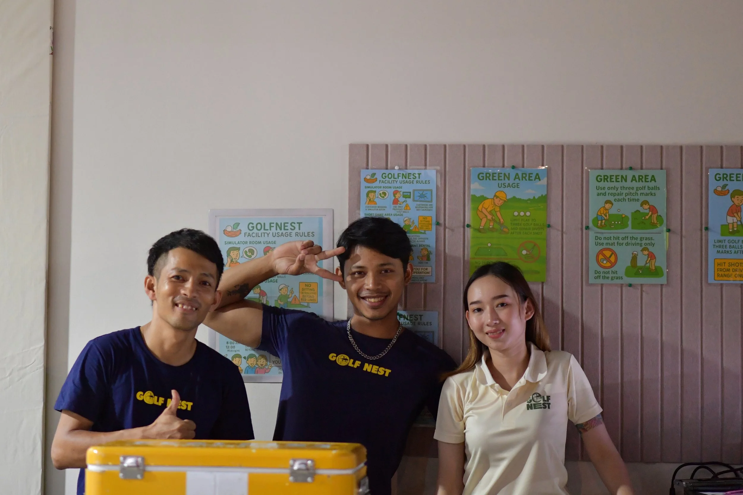 Three smiling staff members at GOLF NEST facility standing in front of signs with facility rules, one person making a peace sign, another giving a thumbs-up.