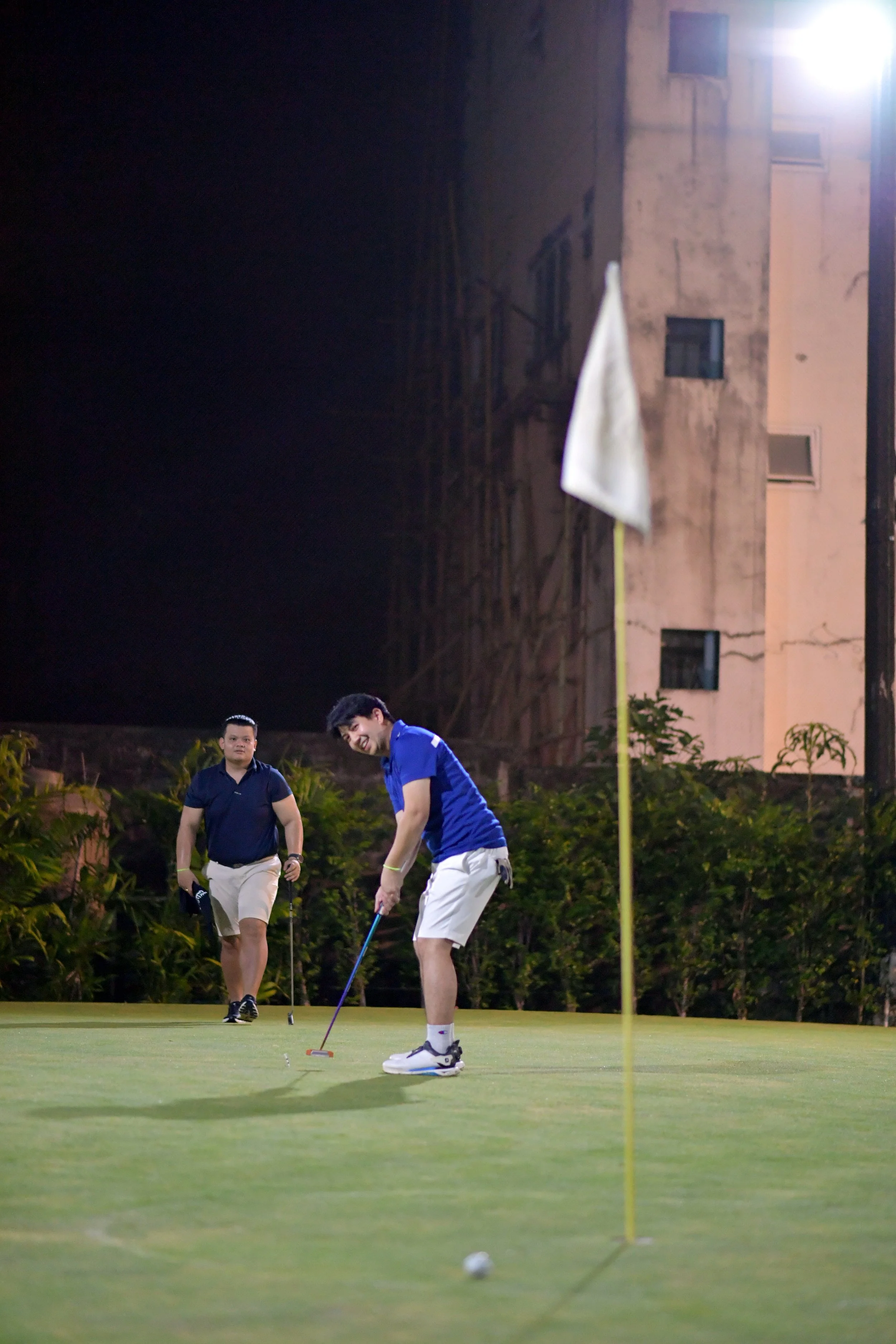 Two men playing golf on a well-lit green field at night, with one about to putt and the other observing, near a white flag.