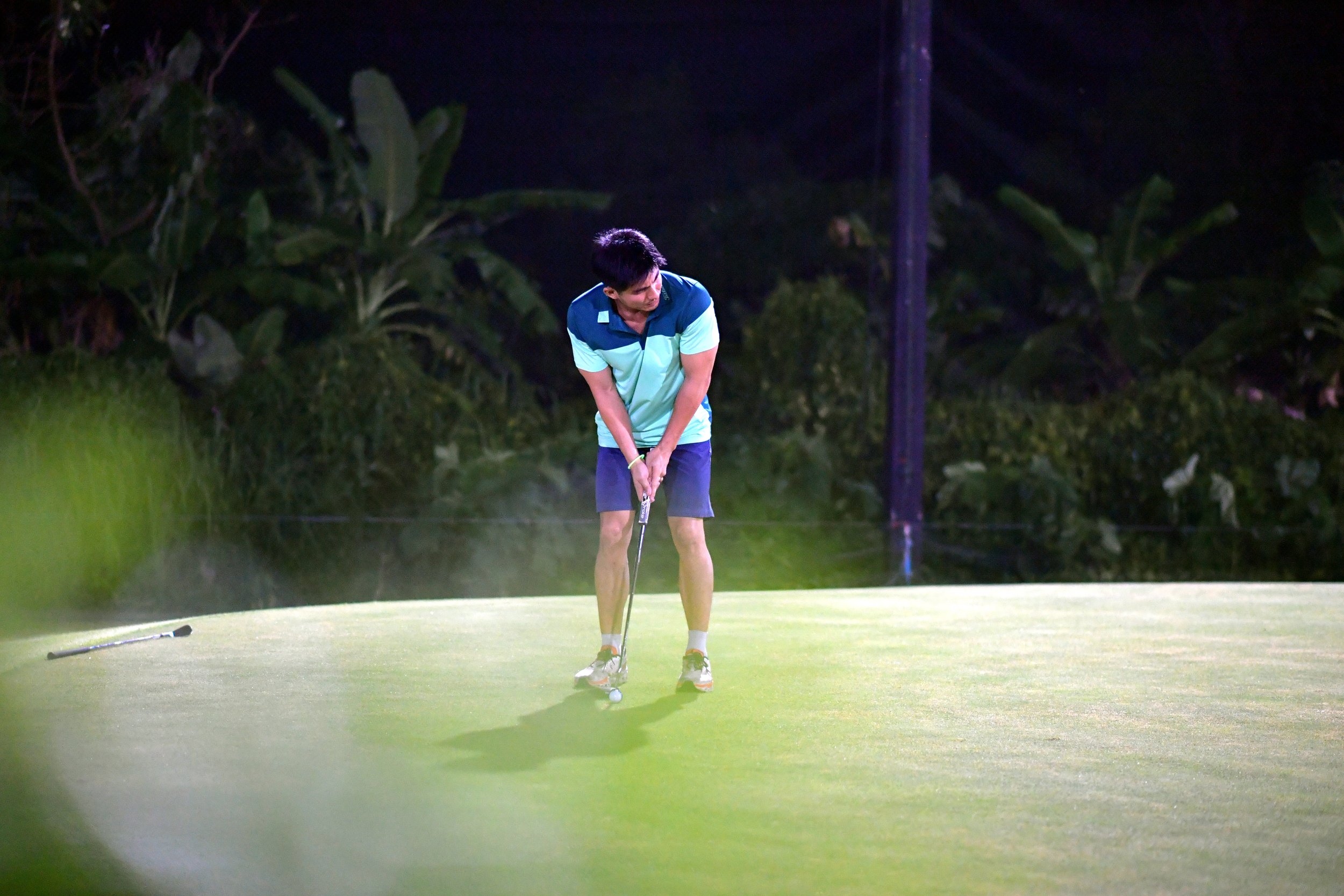 A young man playing golf at night on a bright green putting green, preparing to putt with a golf club, with trees and a utility pole in the background.