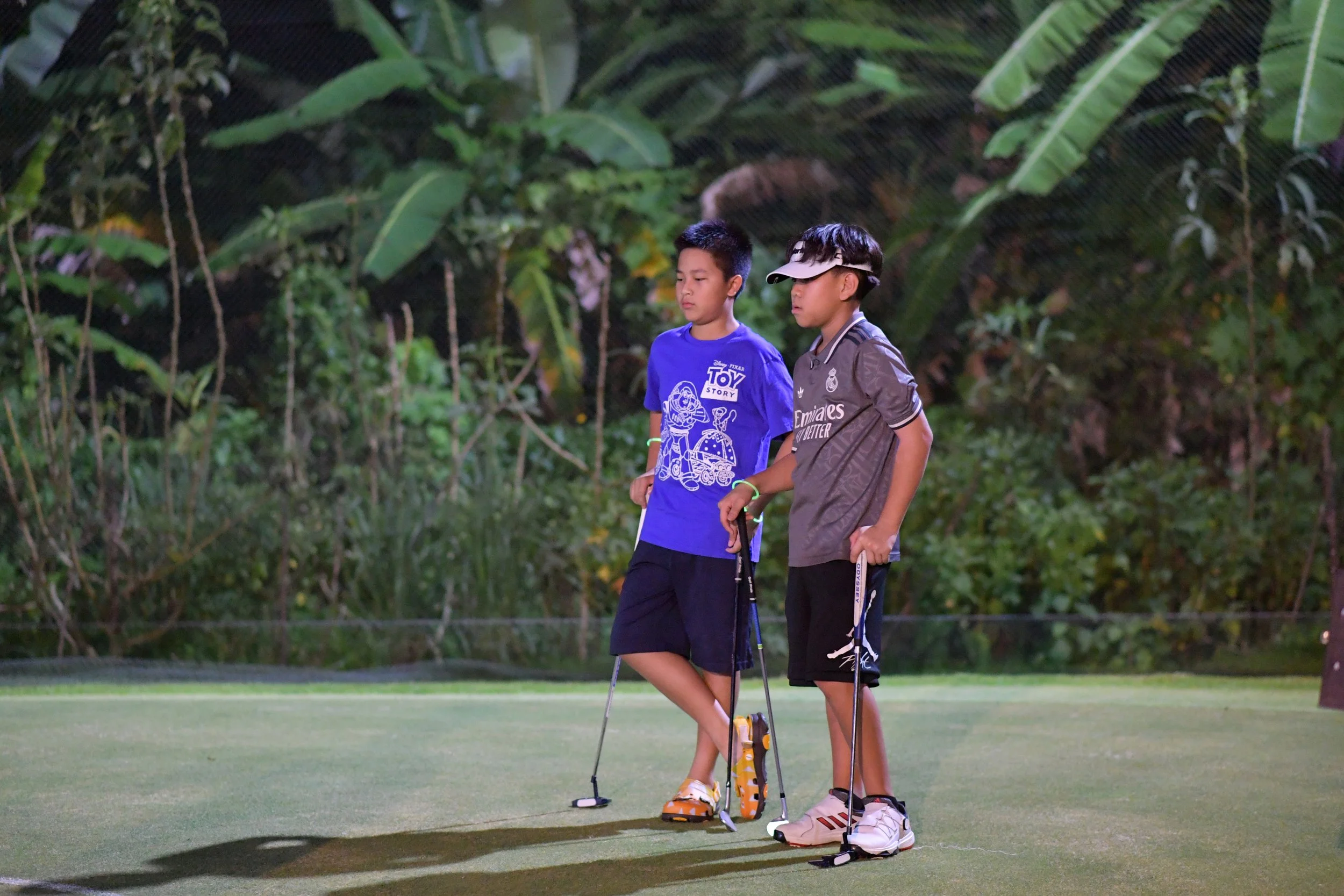 Two boys standing on a golf course holding golf clubs, with a lush green background of tropical plants.