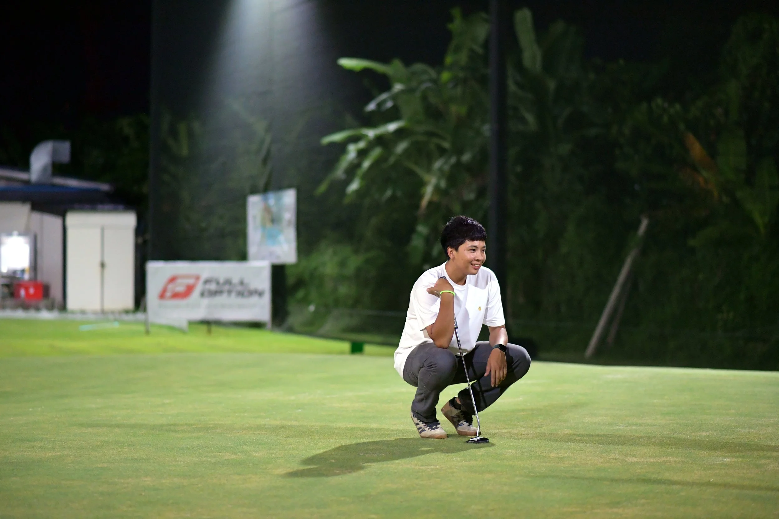 A young man crouching on a golf course at night, holding a golf club, smiling.