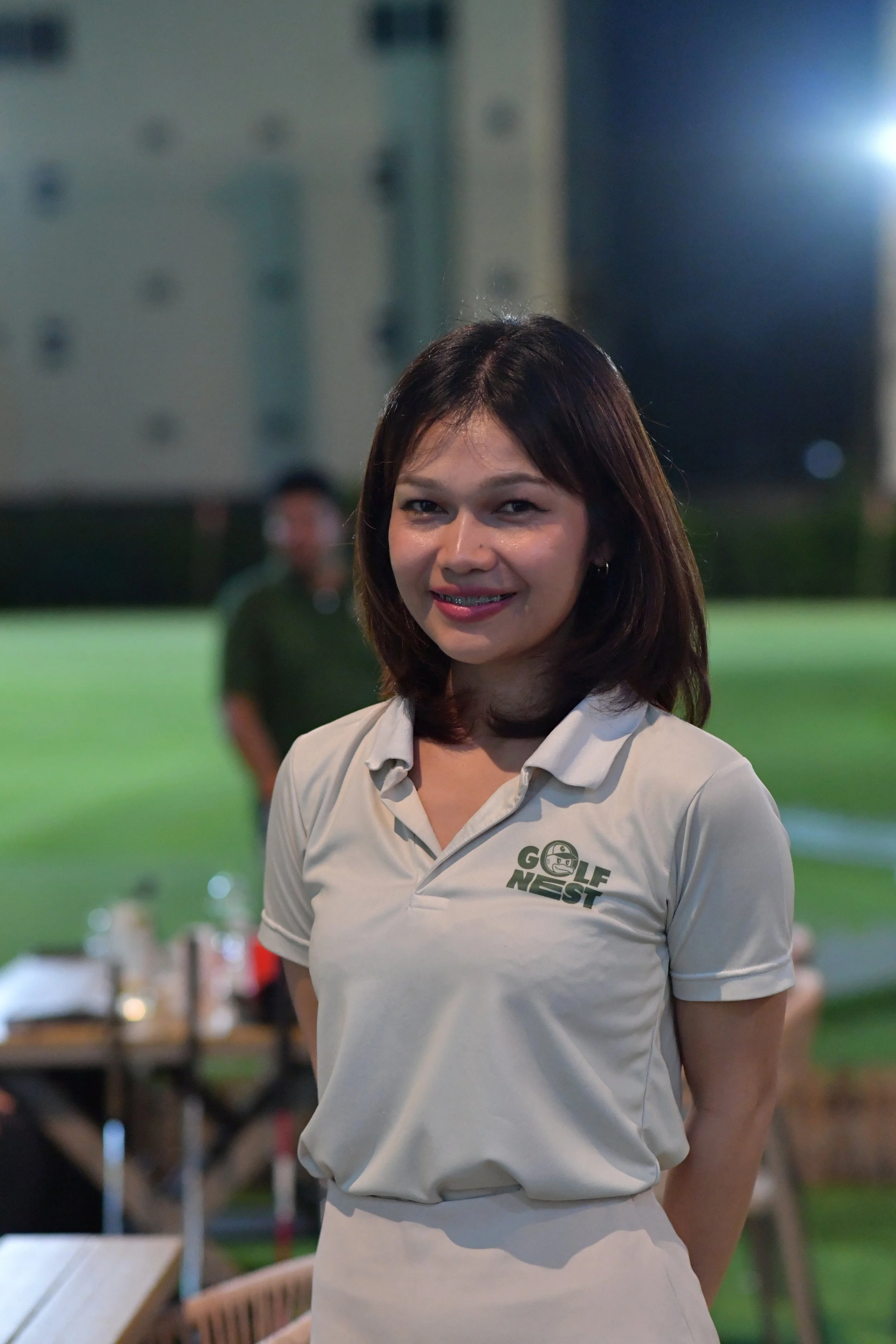 A woman smiling at the camera wearing a Golf NEST shirt at an outdoor evening event.