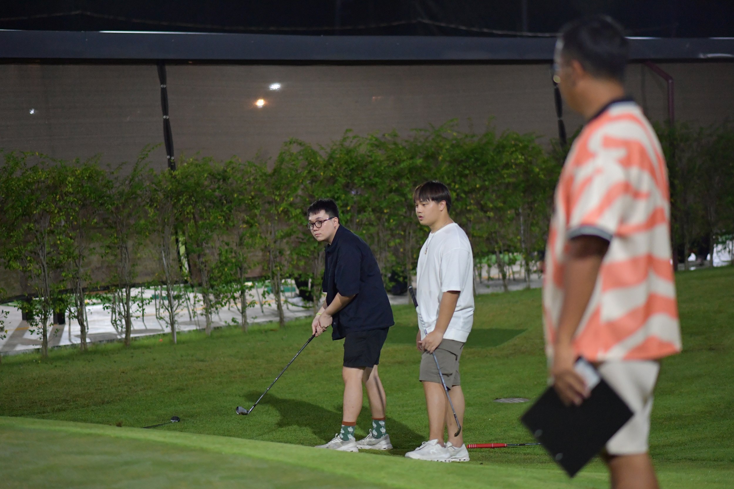 Three young men on a golf course, two holding golf clubs and one observing, with a person in the foreground holding a clipboard.