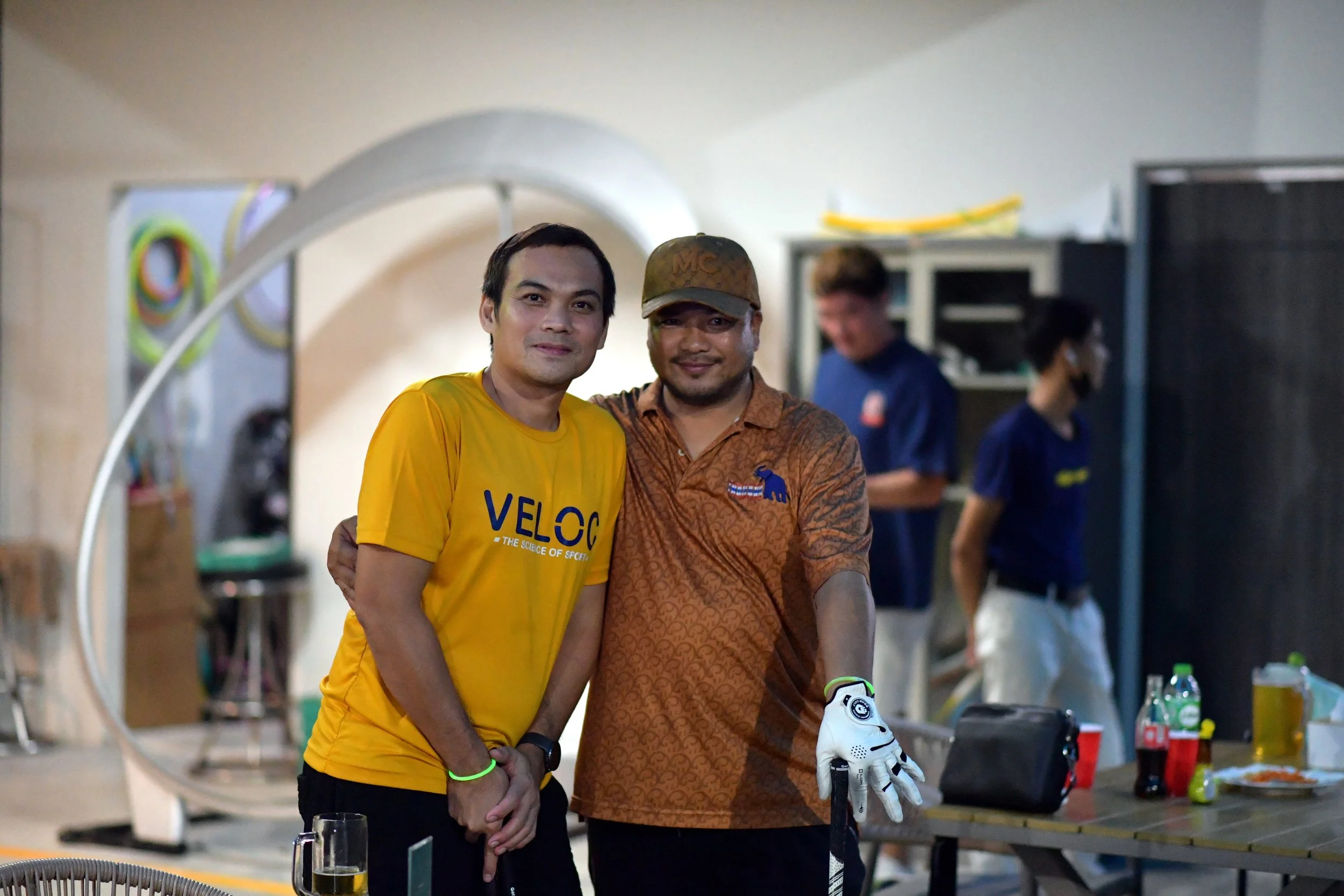 Two men standing together indoors, one wearing a yellow sports shirt and the other in a brown patterned shirt with a cap and a golf glove, posing for a photo.