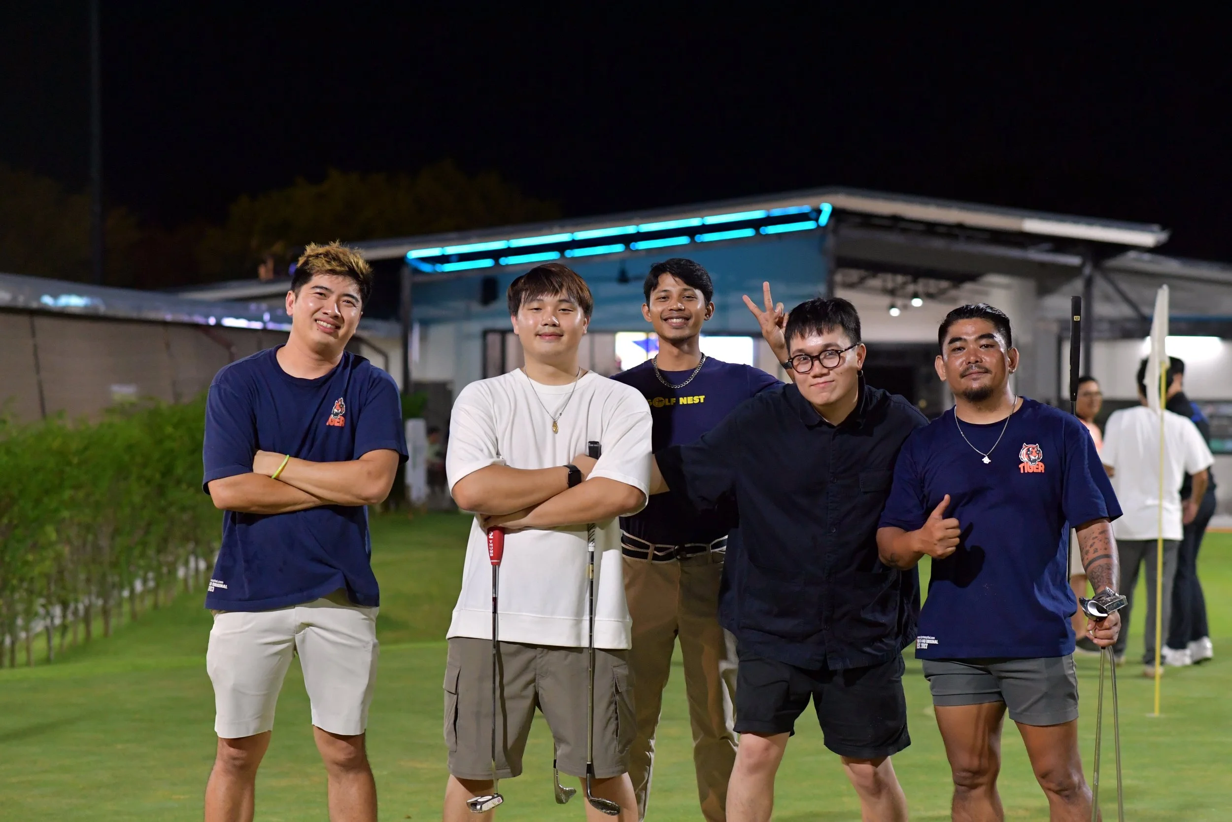 A group of five young men standing on a golf course at night, smiling and posing for the photo, with a building and other people in the background.
