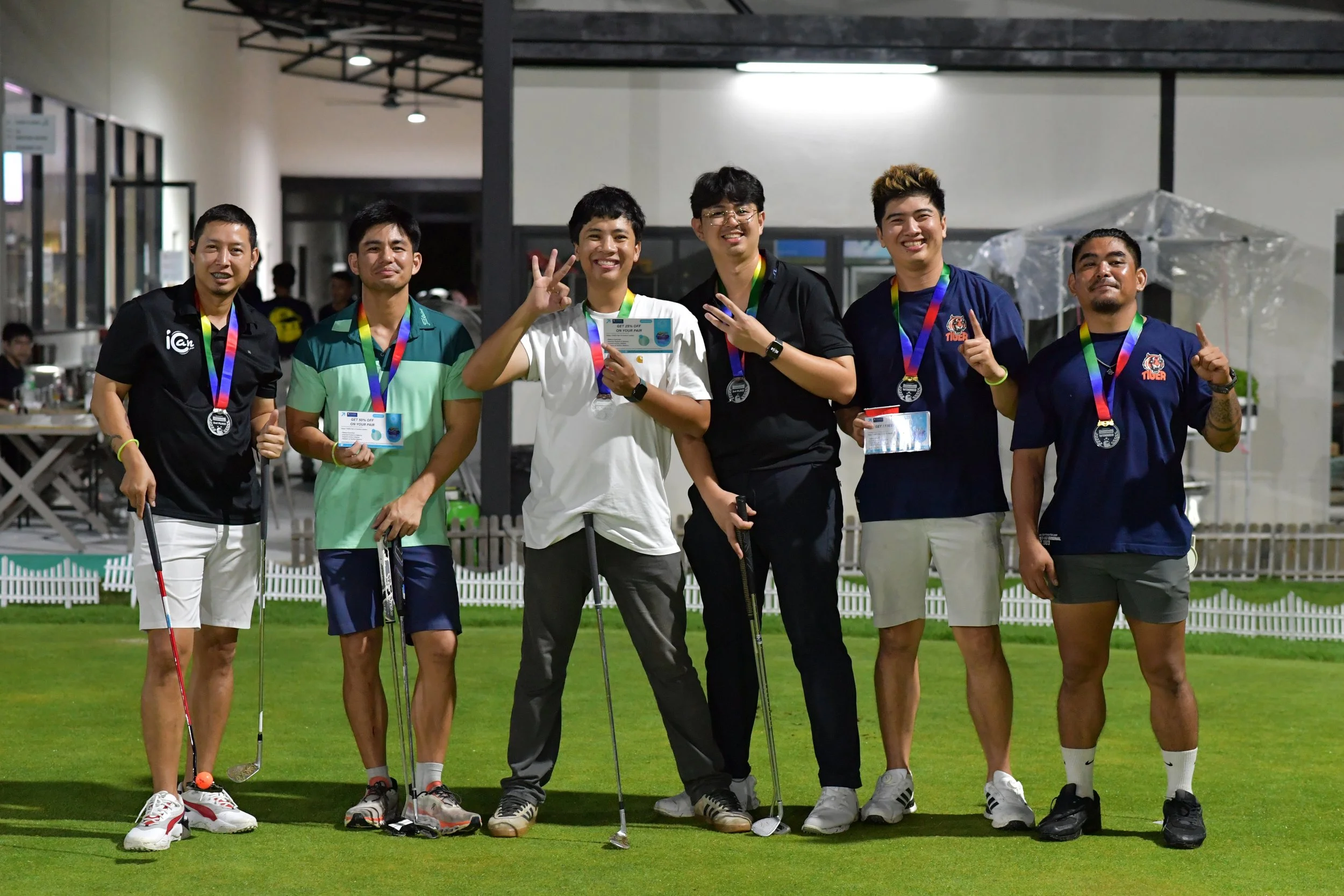 A group of six young men standing on a golf course, smiling and showing medals around their necks. They are holding golf clubs and appear to be celebrating a successful game or tournament.
