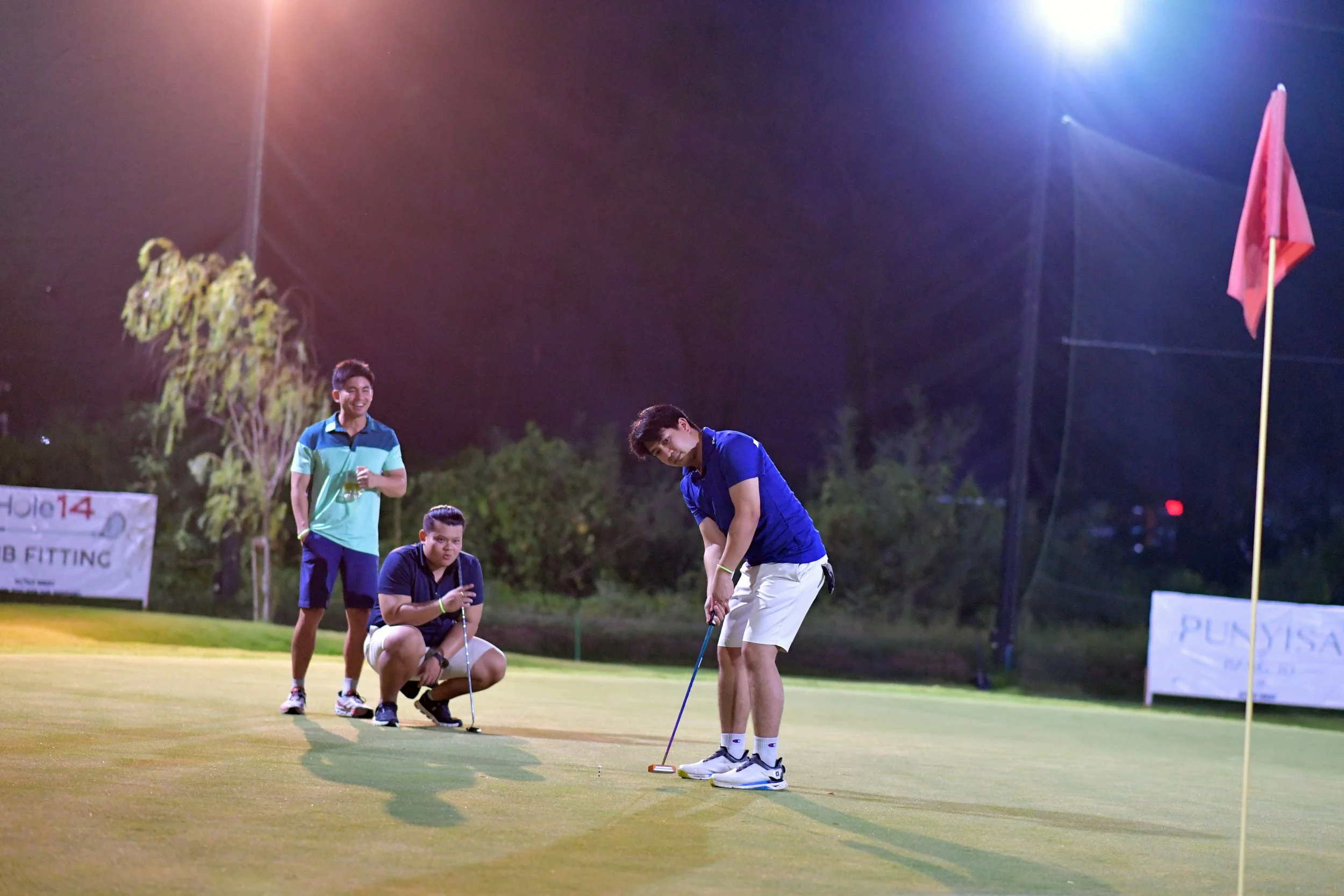 Three young men playing golf on a putting green at night, illuminated by bright lights, with trees and banners in the background.