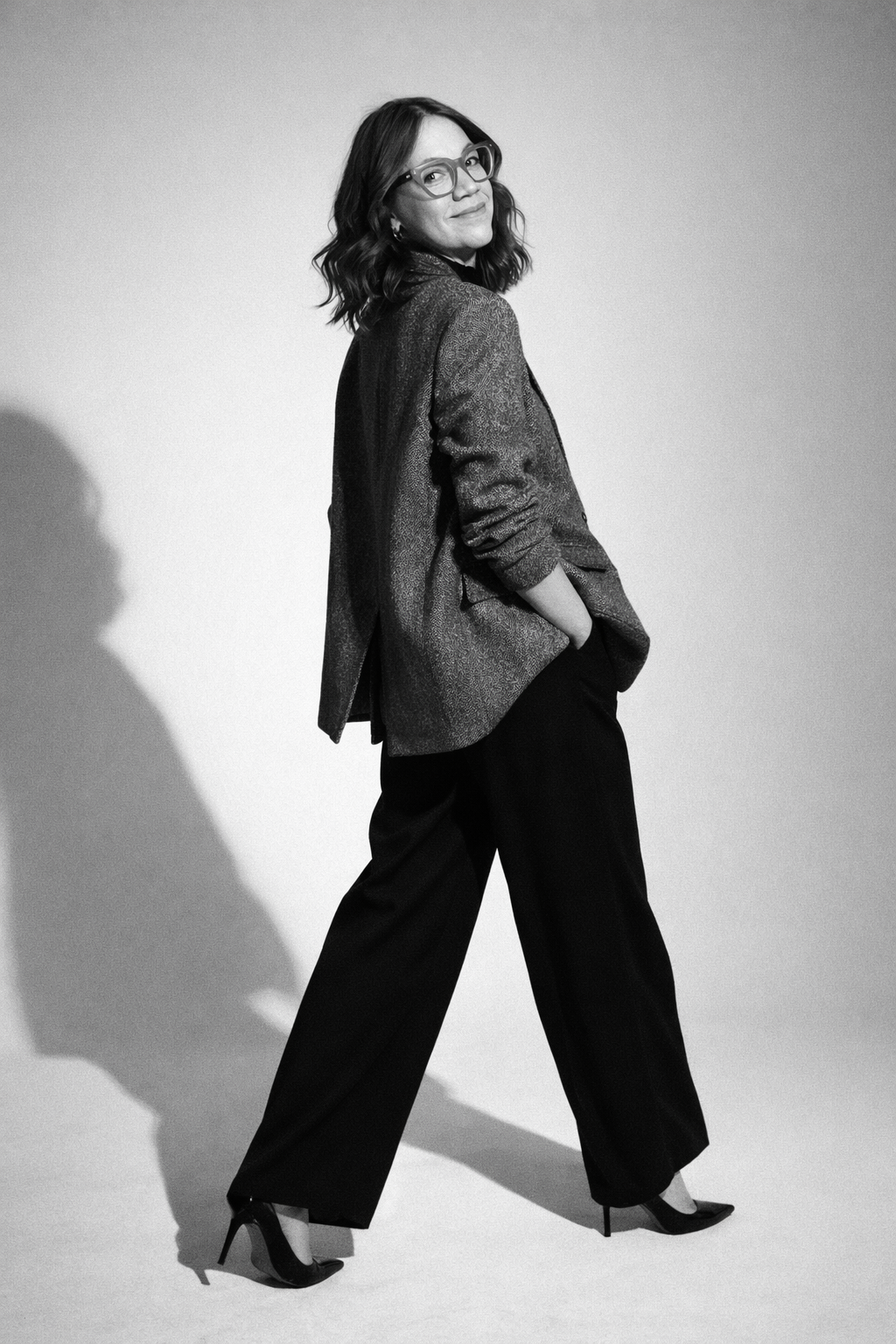 Schwarz-weißes Fashionfoto einer Frau mit braunen Haaren und Brille im oversized Tweed-Blazer, weiter schwarzer Hose und High Heels, dynamische Pose vor minimalistischem Studio-Hintergrund mit weichem Licht und langen Schatten.