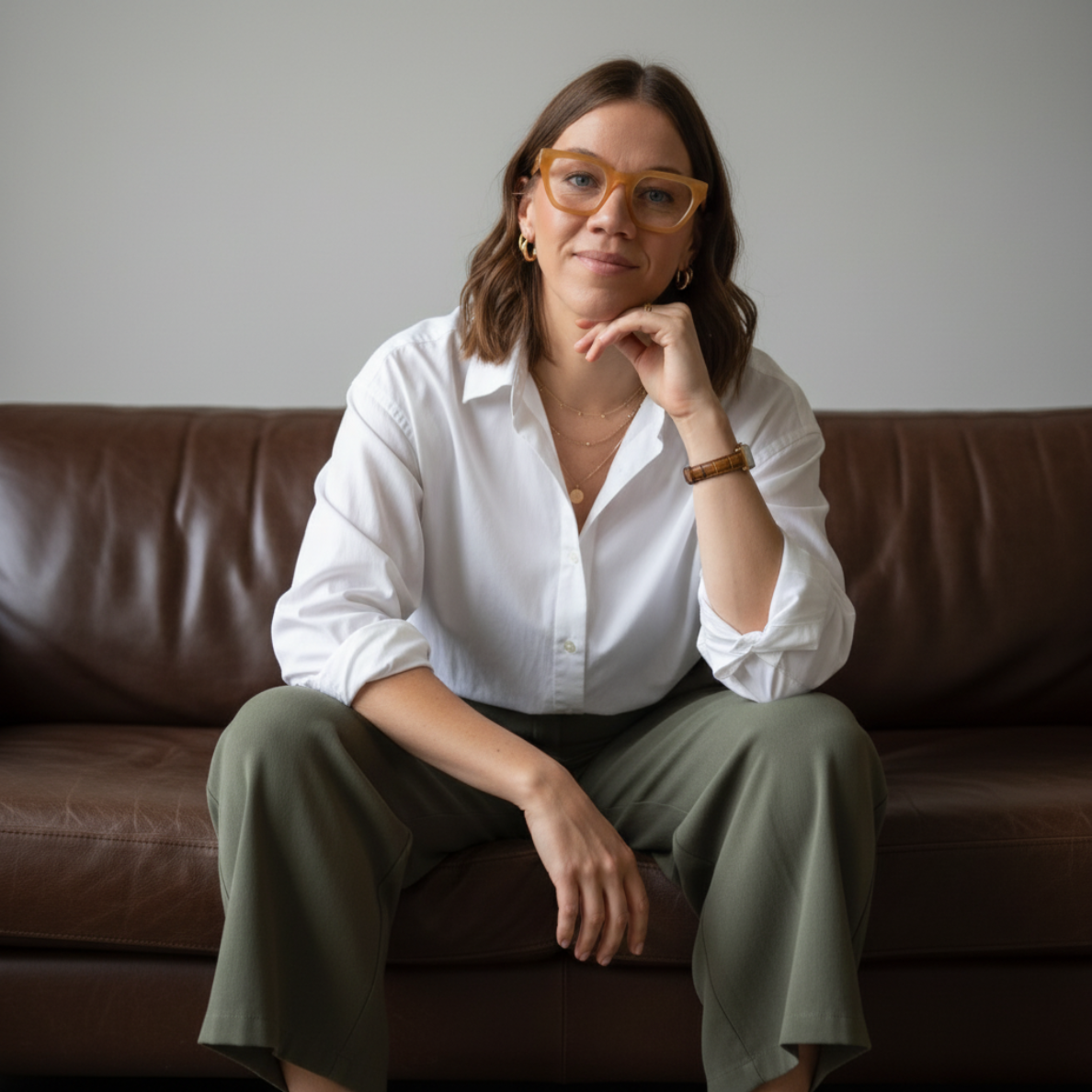 Person sitzt auf einem braunen Ledersofa und blickt in die Kamera. Sie trägt ein weißes Hemd, olivgrüne Hose und eine Brille mit hellem Rahmen. Der Hintergrund ist neutral und hell.