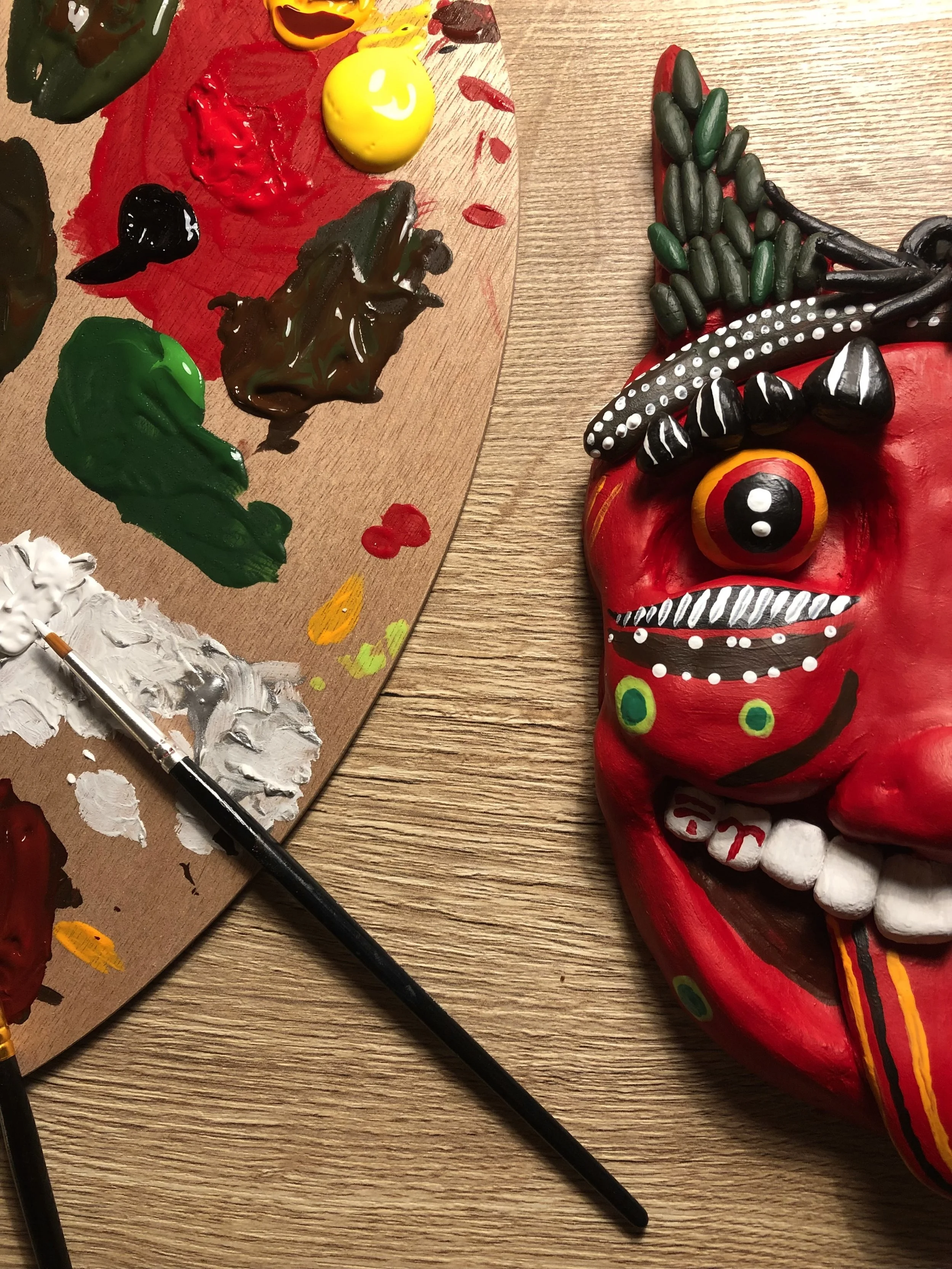 Bemalte Tonmaske in kräftigen Rot-, Grün- und Schwarztönen liegt neben einer Holzpalette mit Acrylfarben und einem Pinsel auf einem Tisch.