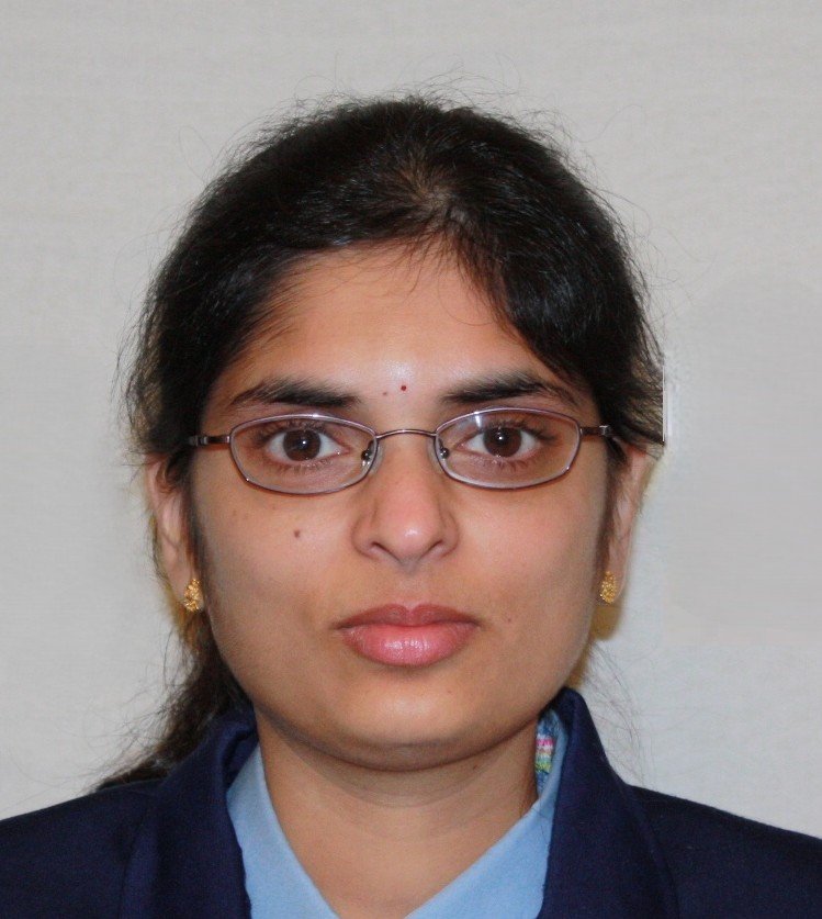 A woman wearing glasses and gold earrings, dressed in a dark blazer and light blue shirt, against a plain background.