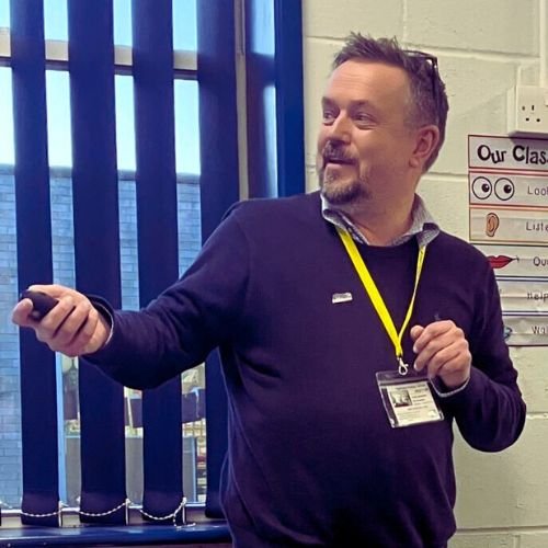 A man in a dark sweater with a yellow lanyard and identification badge, standing in a classroom, holding a remote control or pointer, with blue vertical window blinds behind him.