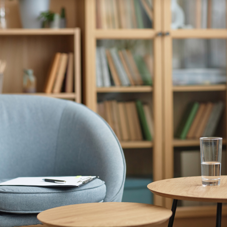 Living room with a blue armchair, a clipboard with a pen resting on it, a wooden side table with a glass of water, and a bookshelf filled with books in the background.