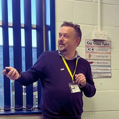 A man giving a presentation in a classroom, holding a remote, with class rules posted on the wall behind him.