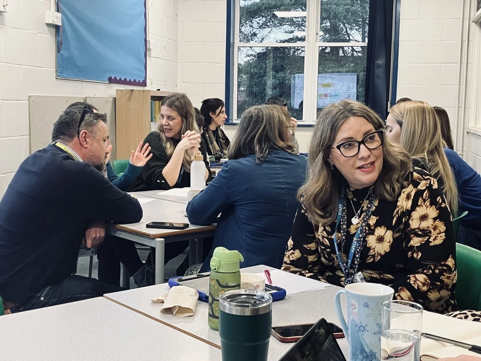 Group of adults sitting at tables in a classroom or meeting room, engaged in conversation. Some are talking while others listen.